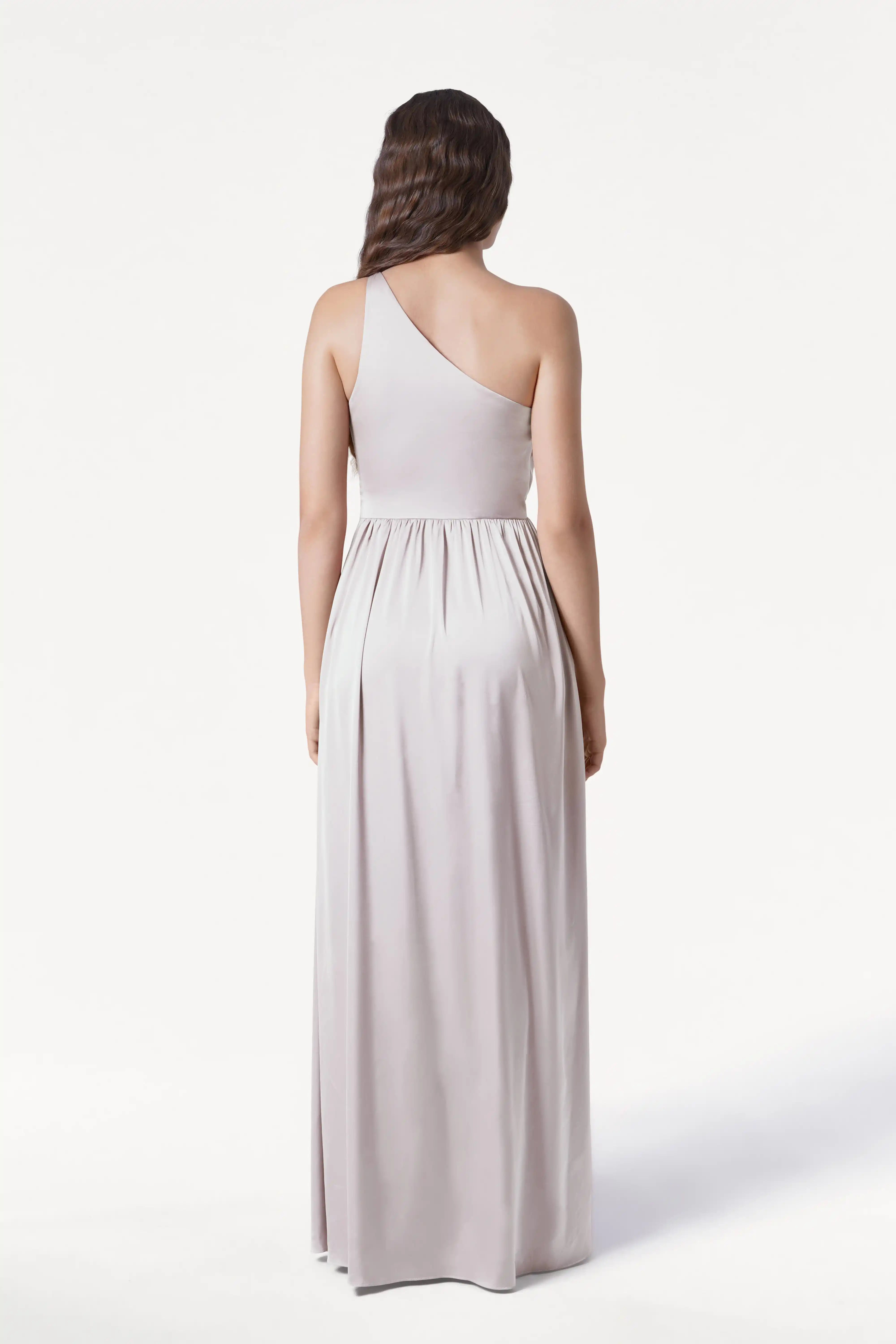 A woman with long, wavy brown hair faces away, wearing the TH&TH Greta Bridesmaid Dress in Oyster Satin—a floor-length, one-shoulder gown with Grecian draping—set against a plain white background.