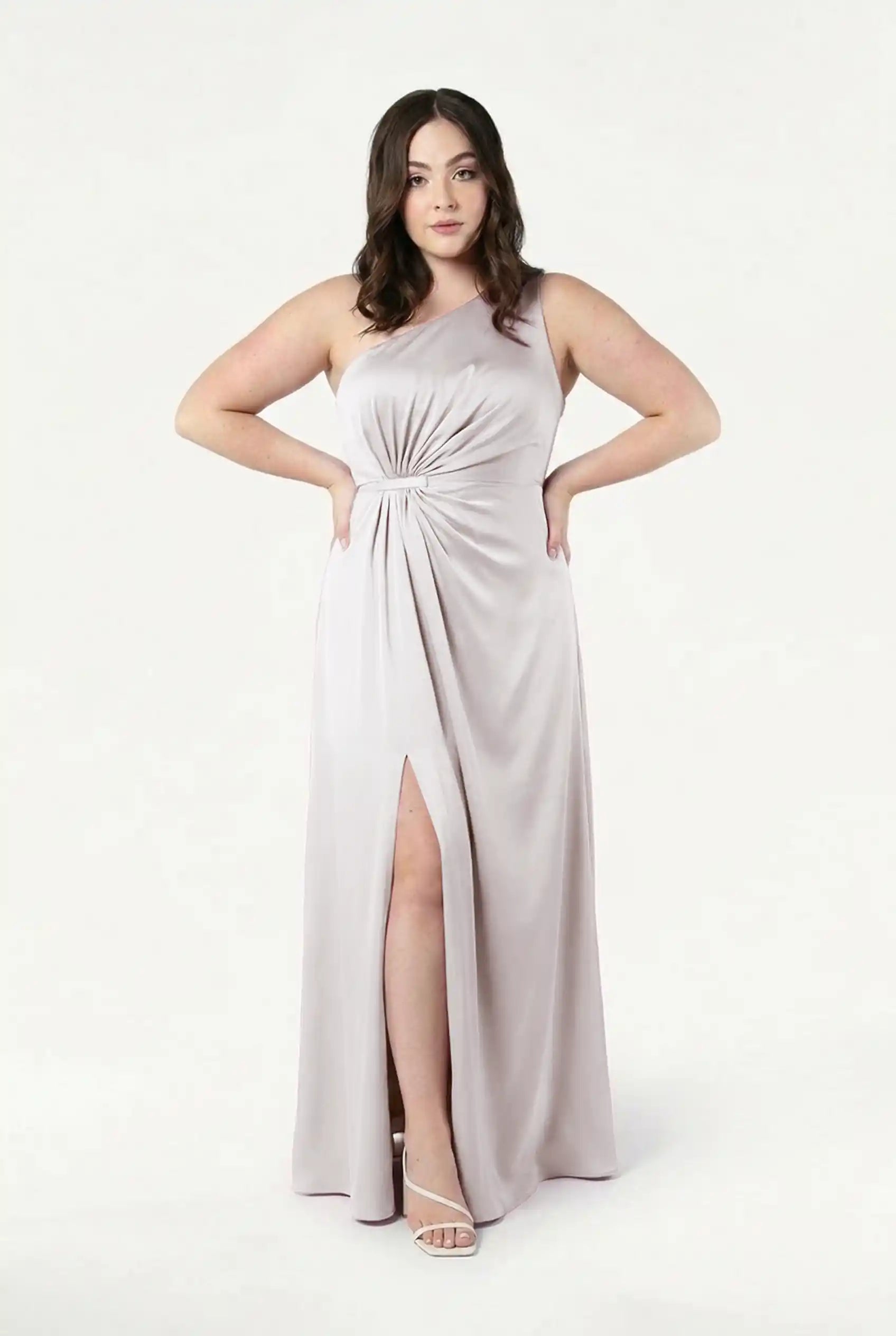A woman faces forward in the Greta Bridesmaid Dress in Oyster Satin by TH&TH, featuring a one-shoulder Grecian drape, gathered waist, and front slit. She wears strappy white heels with neutral makeup and medium-length dark hair against a plain background.