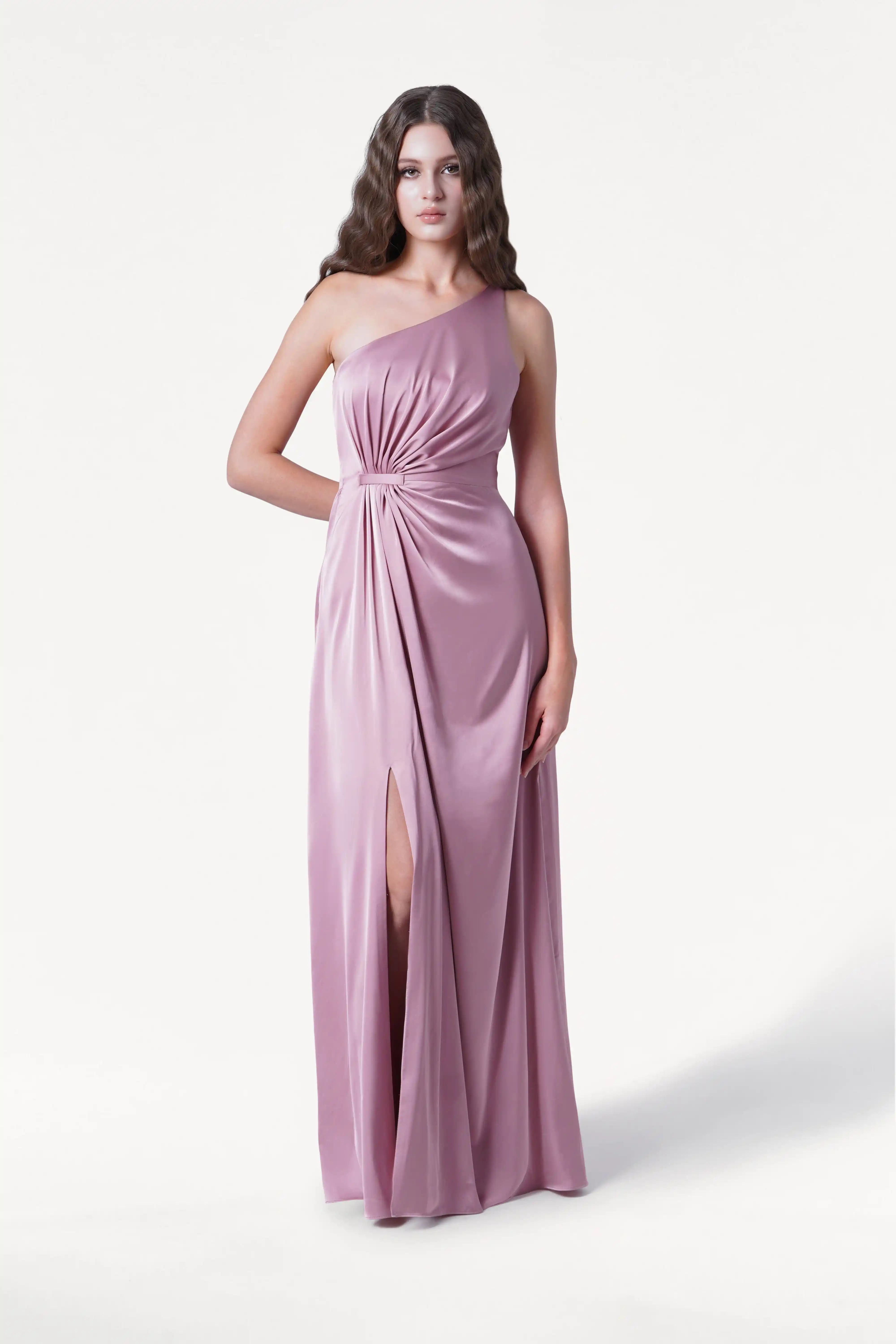 A person wearing the TH&TH Greta Bridesmaid Dress in rose pink satin, featuring a sleeveless design, Grecian draping, gathered waist, and high slit, stands against a plain white background.