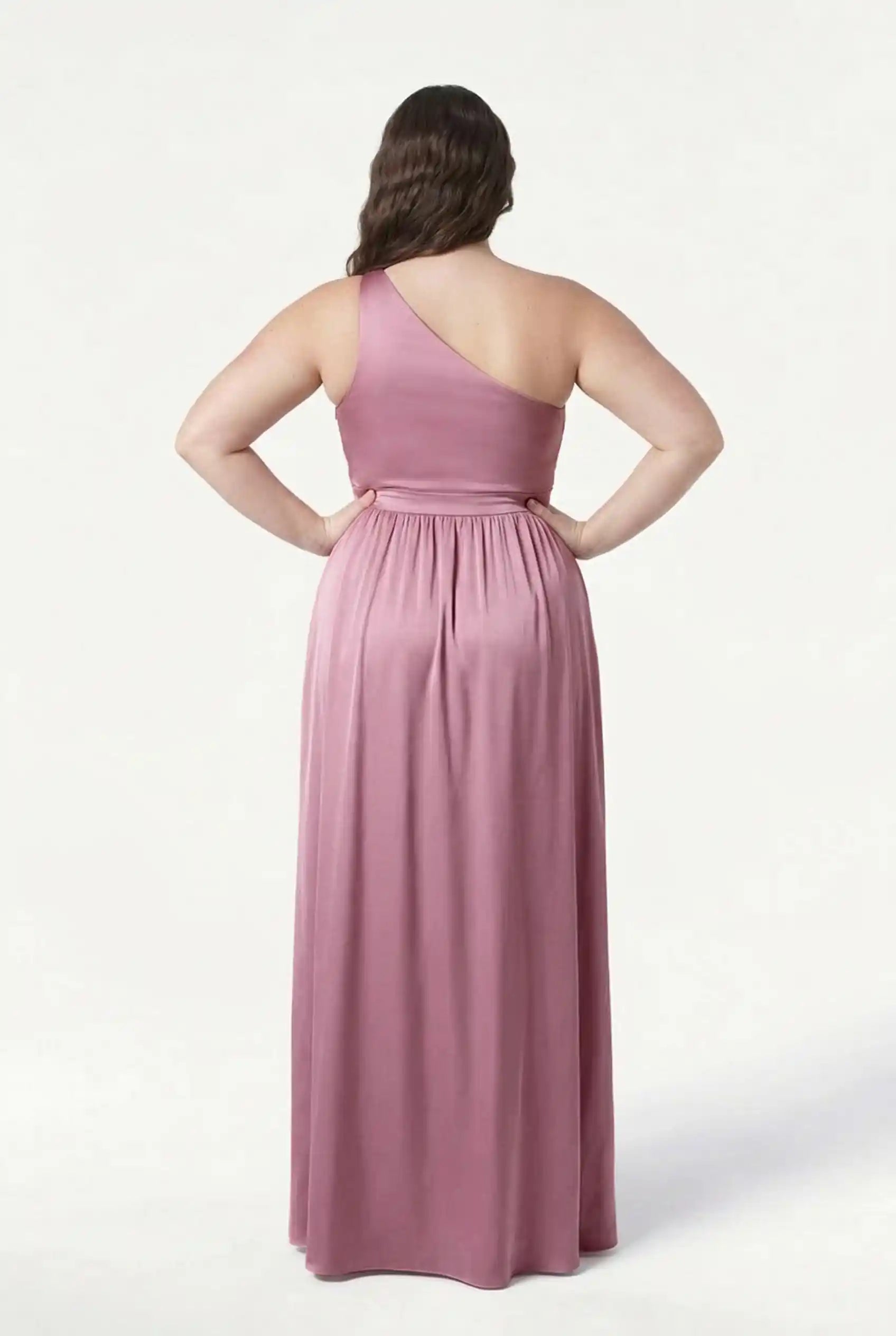 A woman stands facing away, wearing the TH&TH Greta Bridesmaid Dress in Rose Pink Satin—a long, sleeveless one-shoulder gown with Grecian draping and a gathered waist, elegantly flowing to the floor against a plain white background.