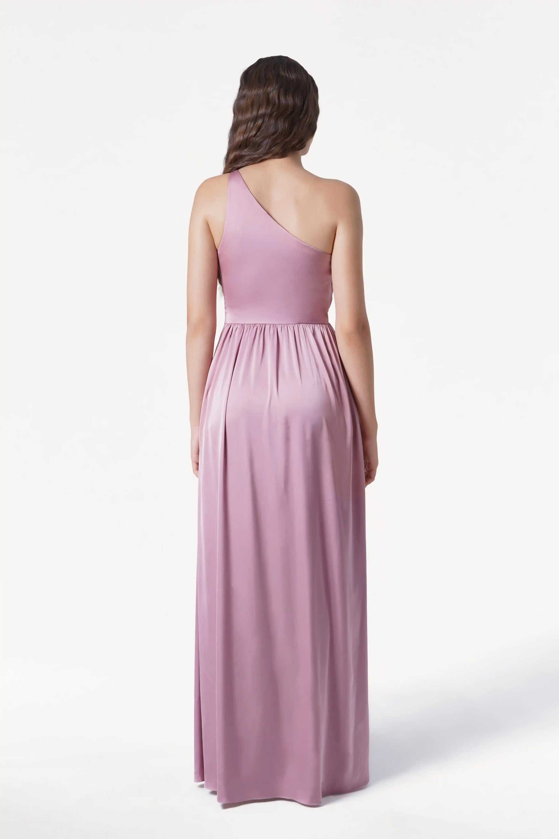 A woman faces away in the Greta Bridesmaid Dress by TH&TH, a long rose pink satin one-shoulder gown with Grecian draping and a gathered waist. Her long wavy hair contrasts against the plain white background.