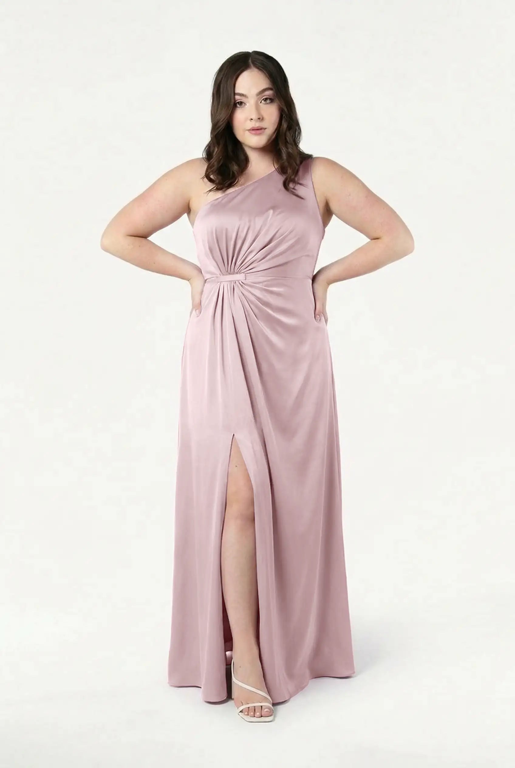 A woman faces forward against a white background in the TH&TH Greta Bridesmaid Dress in Smoked Blush Satin—a sleeveless, floor-length gown with a knotted waist and thigh-high slit—paired with white strappy heels.