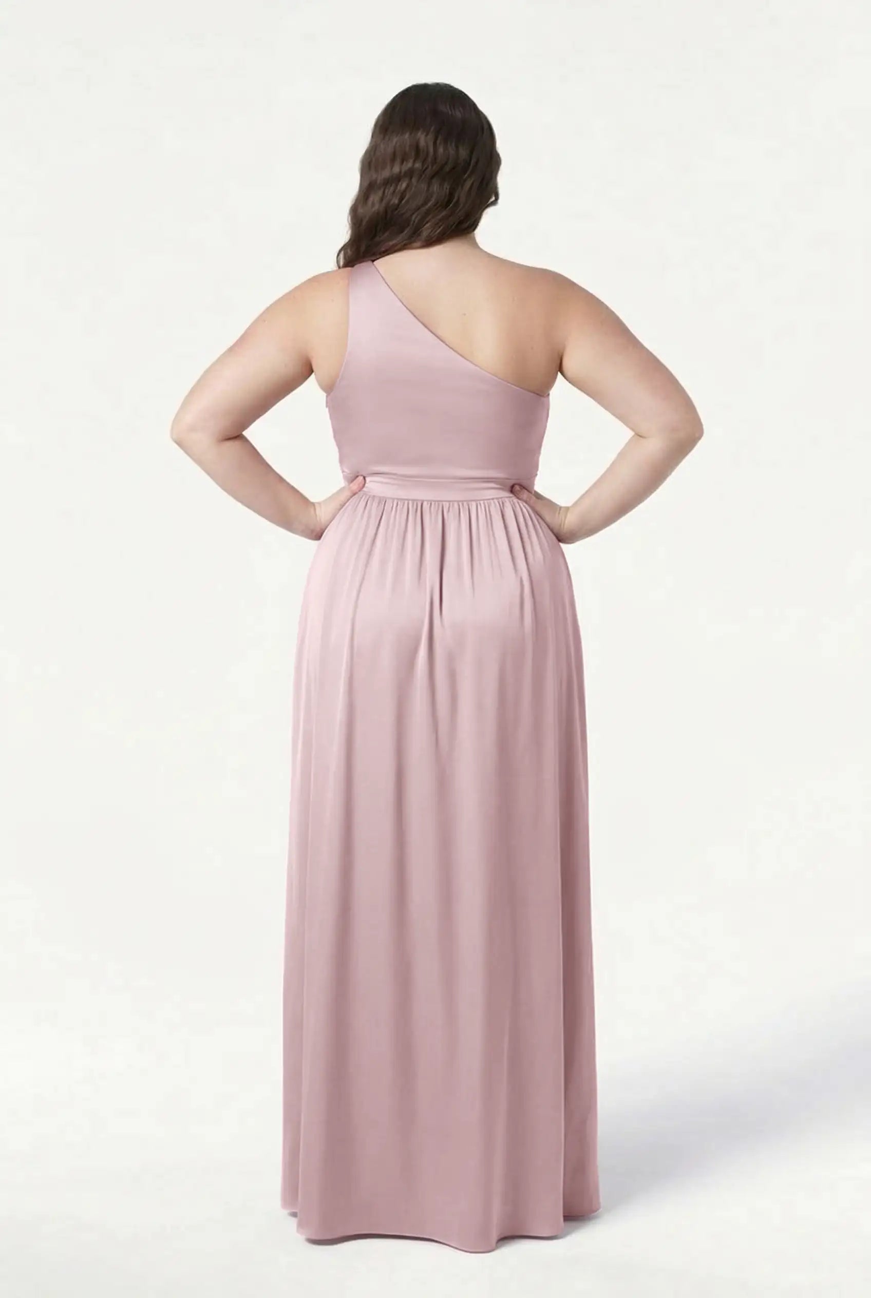 A person with long, wavy brown hair faces away in the TH&TH Greta Bridesmaid Dress—smoked blush satin, floor-length, one-shoulder with Grecian side draping and a gathered waistband—set against a plain white background.