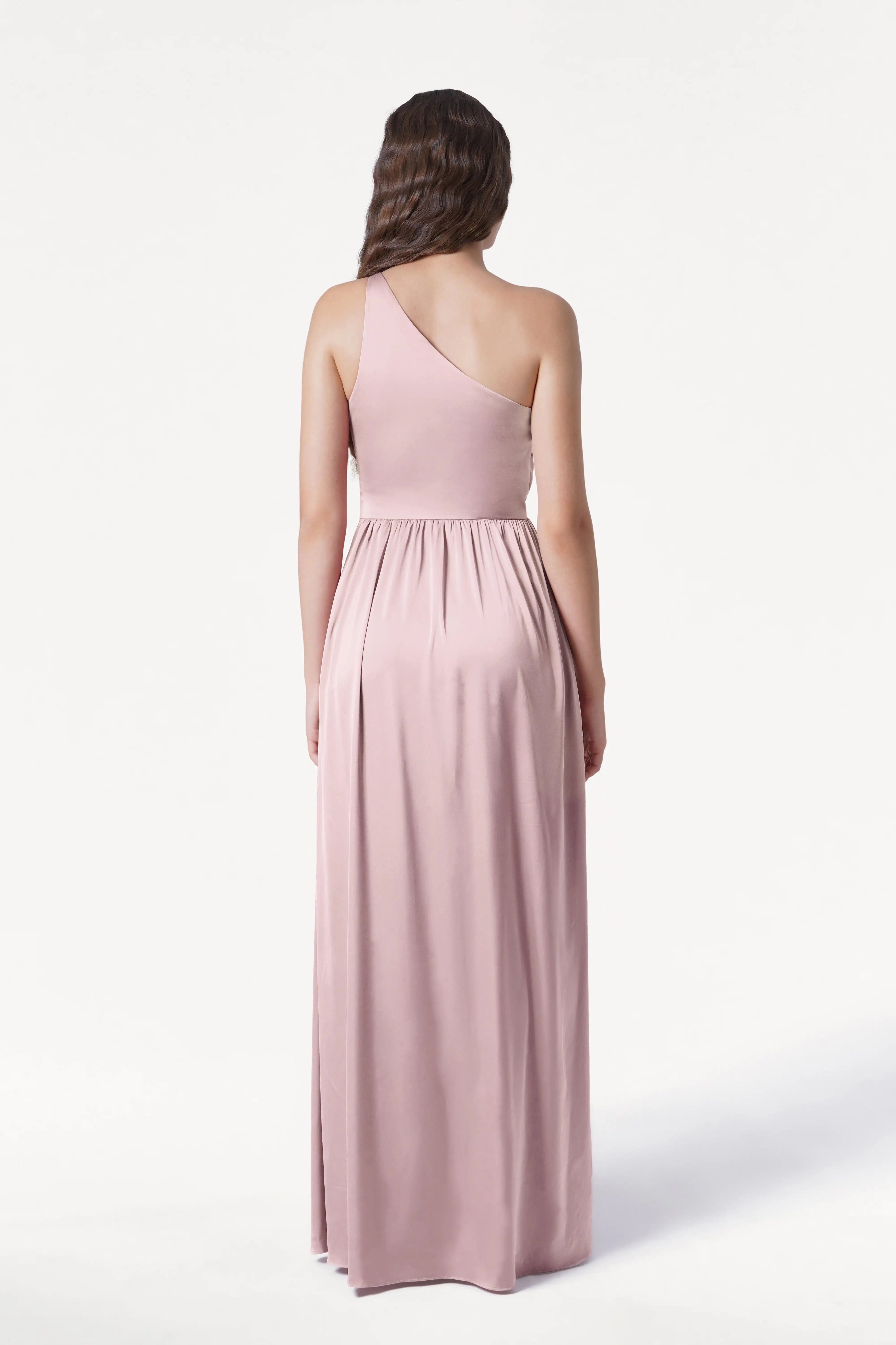 A woman with long brown hair in loose waves stands with her back to the camera, wearing the TH&TH Greta Bridesmaid Dress in Smoked Blush Satin—a floor-length, one-shoulder gown with Grecian draping—against a plain white background.