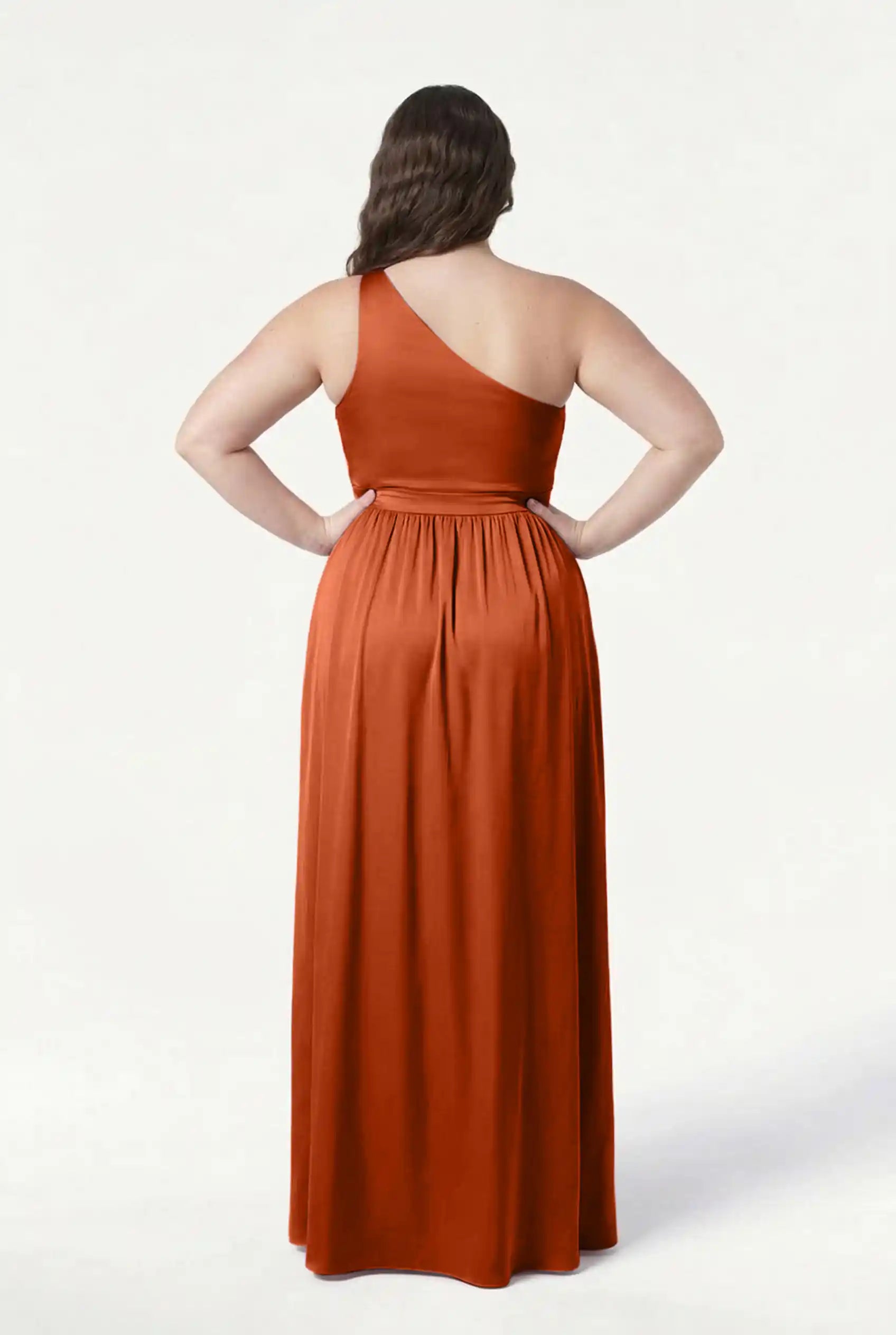 A woman stands with her back to the camera, wearing the TH&TH Greta Bridesmaid Dress in Terracotta Satin, a floor-length gown featuring Grecian draping and a gathered waist. Her hands rest on her hips; dark hair falls to her upper back.