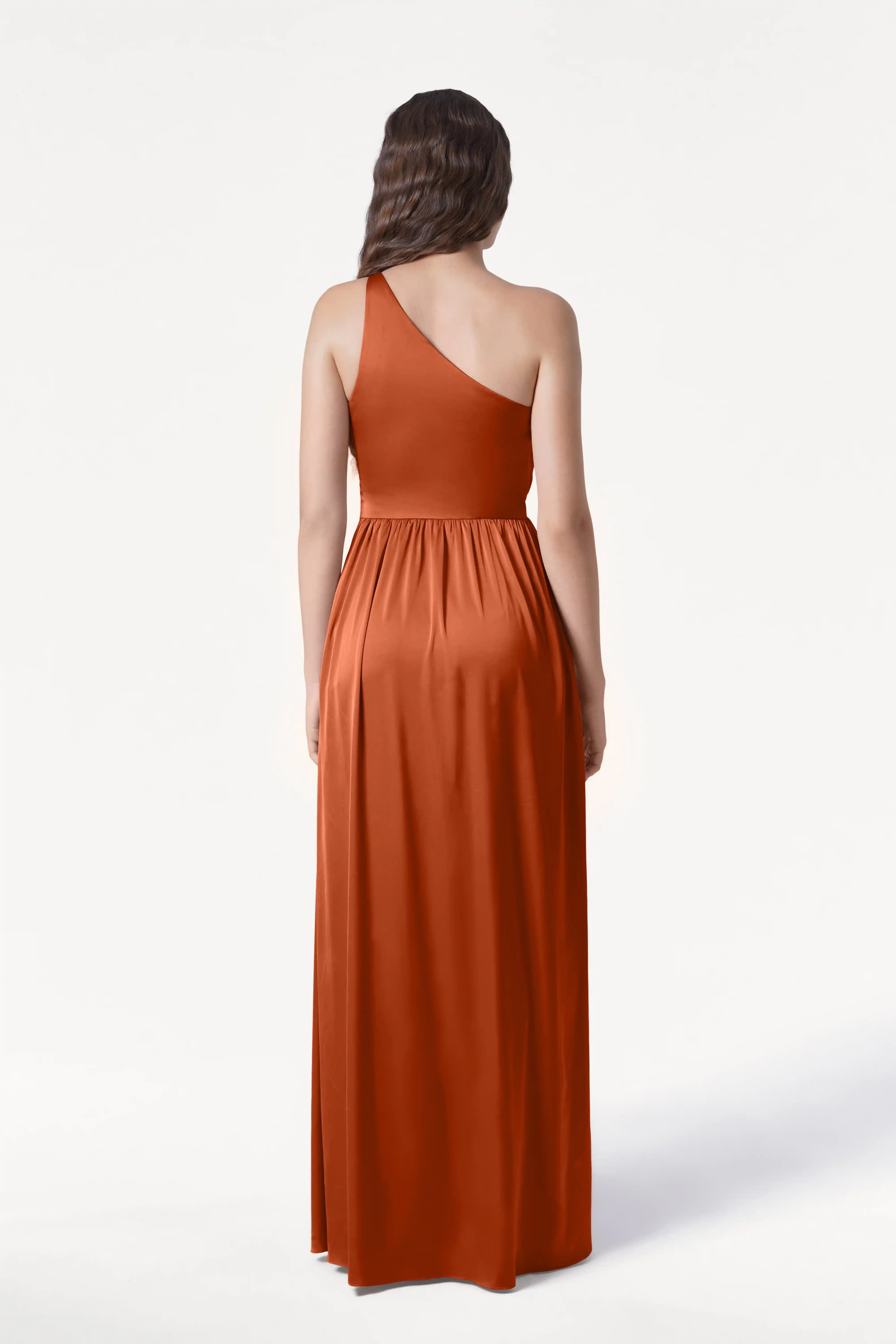 A woman with long, wavy brown hair is seen from the back wearing the TH&TH Greta Bridesmaid Dress in Terracotta Satin—a floor-length, sleeveless one-shoulder gown with Grecian draping—against a plain white background.