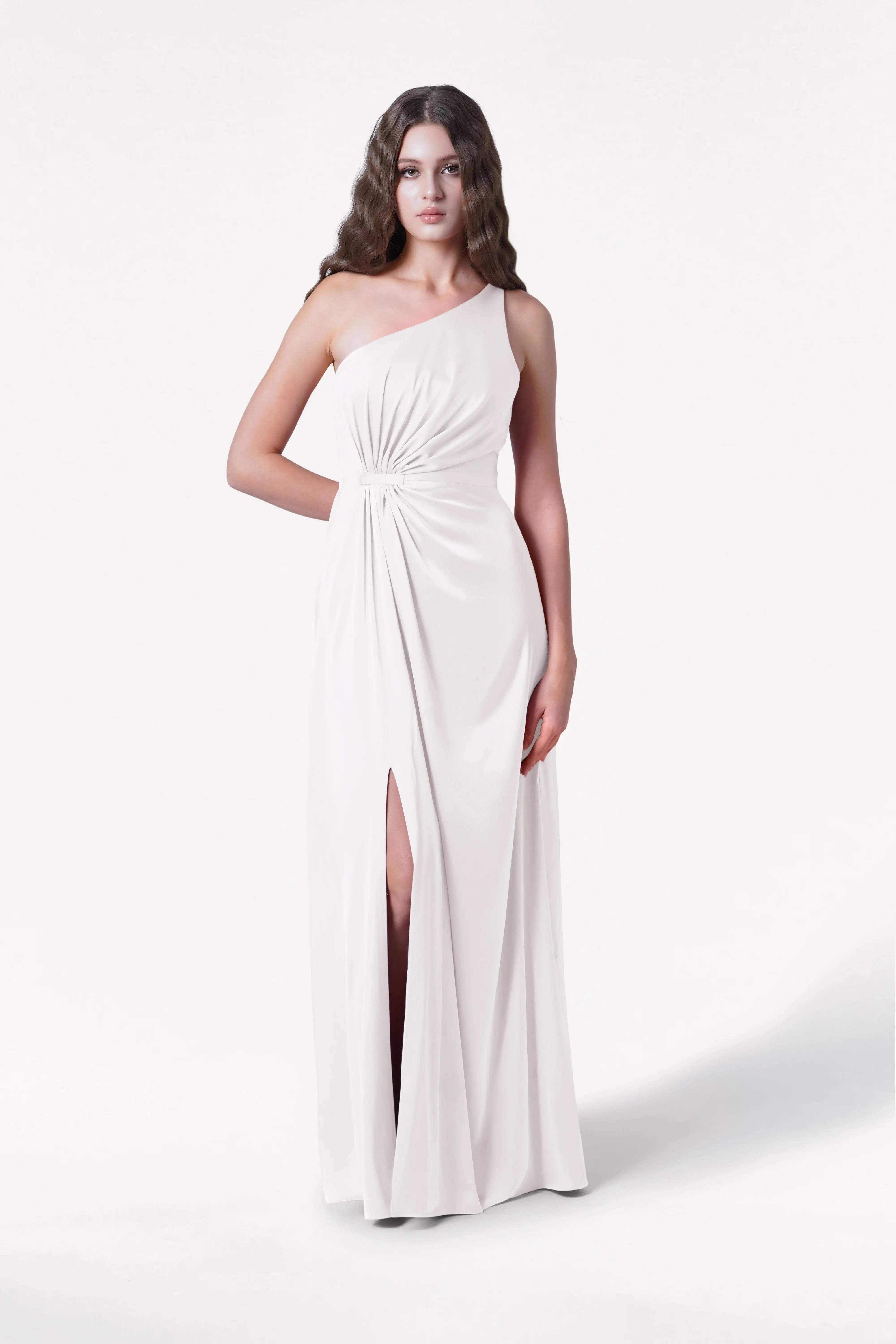 A woman with long wavy brown hair and a neutral expression wears the Greta Dress in Ivory Satin by TH&TH—a long, elegant, one-shoulder gown with a gathered waist and high slit—standing against a white background.