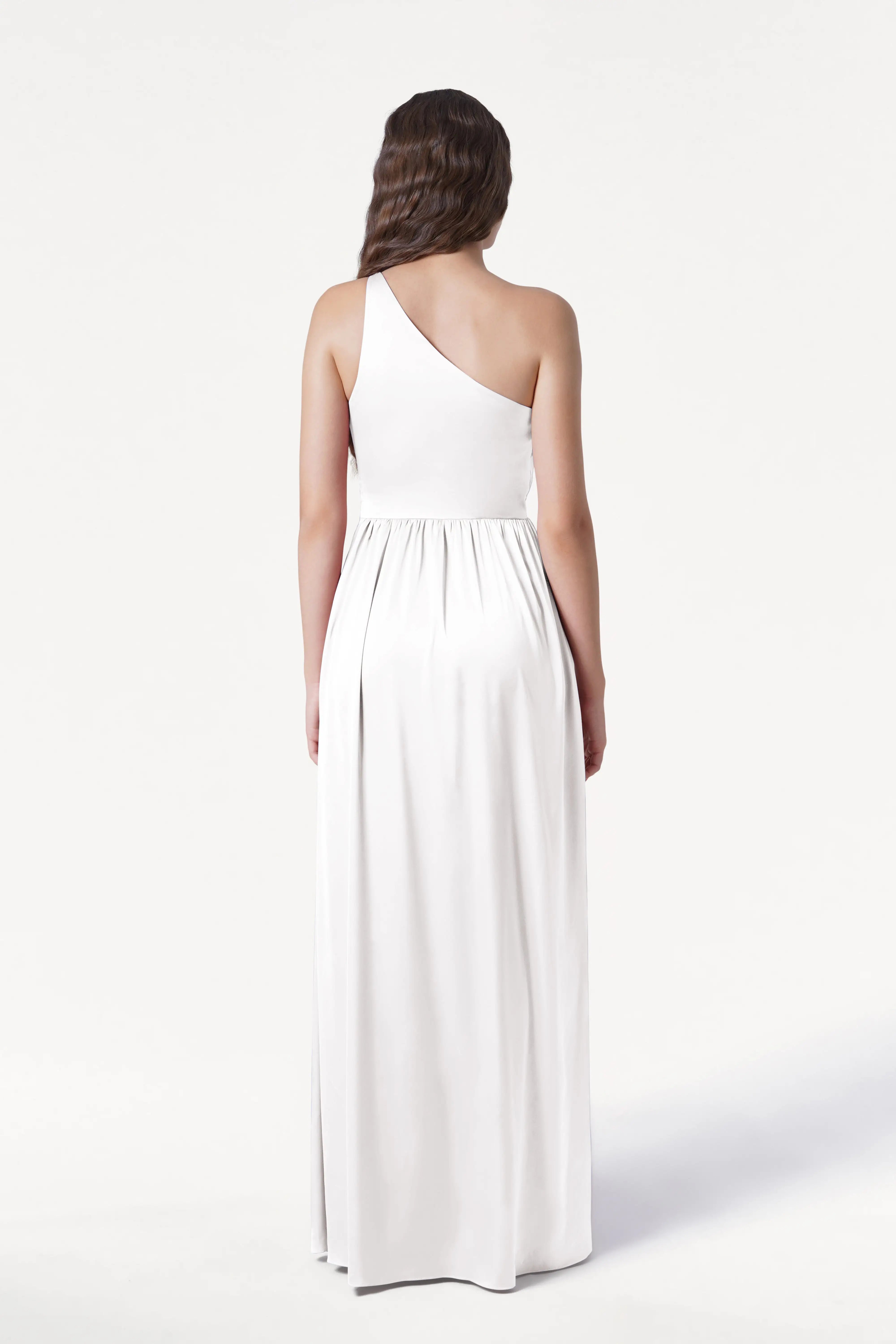 A woman with loose, wavy brown hair stands facing away in the TH&TH Greta Dress in Ivory Satin—a long, elegant, one-shoulder gown—against a plain white background.