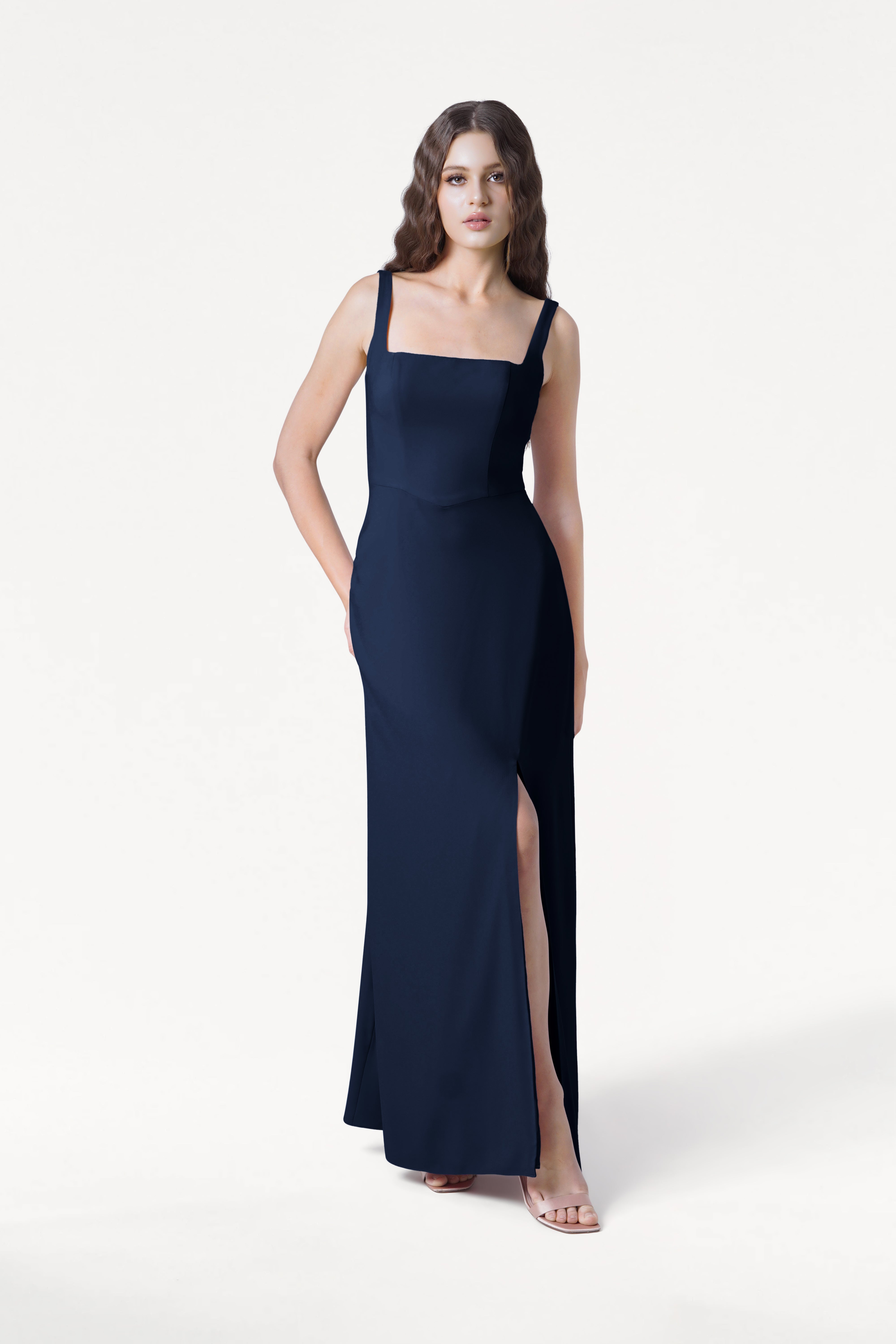 A woman with long wavy brown hair models the TH&TH Helena Bridesmaid Dress in Navy, featuring a sleeveless design, square neckline, and high slit, standing against a plain white background.