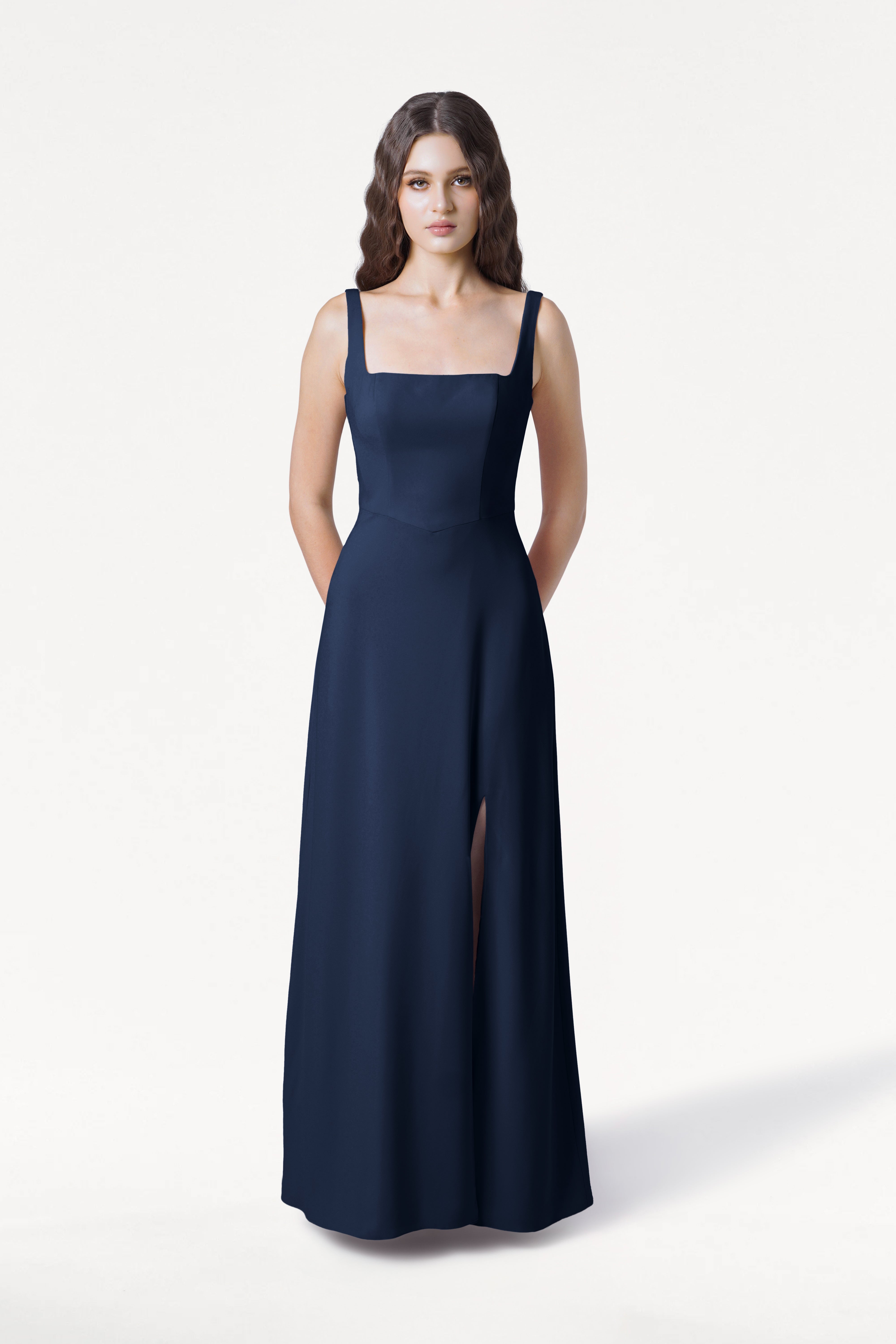 A woman with long wavy hair stands against a plain white background, wearing the TH&TH Helena Bridesmaid Dress in Navy, which features a sleeveless moss crepe design, square neckline, and high slit. Her hands are behind her back.