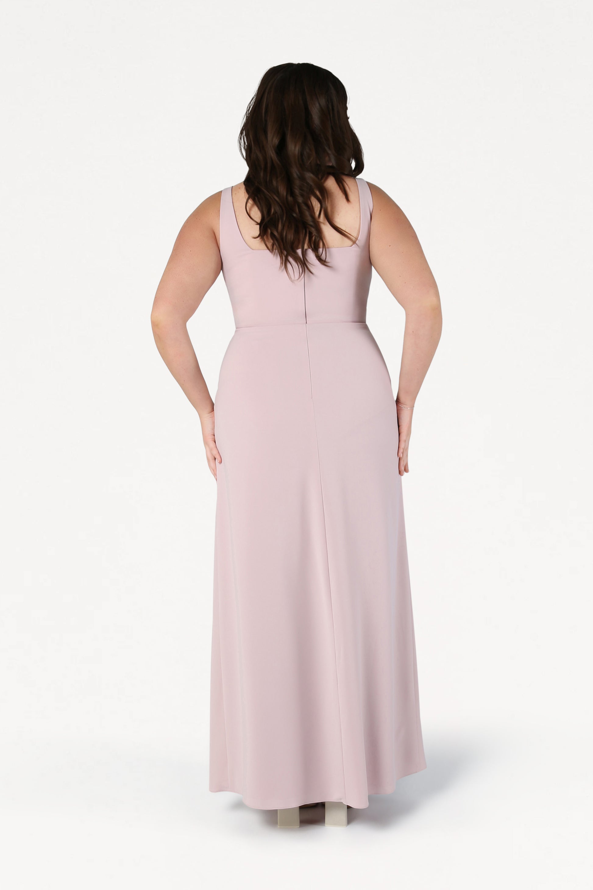A woman with long brown hair stands with her back to the camera, wearing the TH&TH Helena Bridesmaid Dress in Smoked Blush and light-colored shoes against a white background.