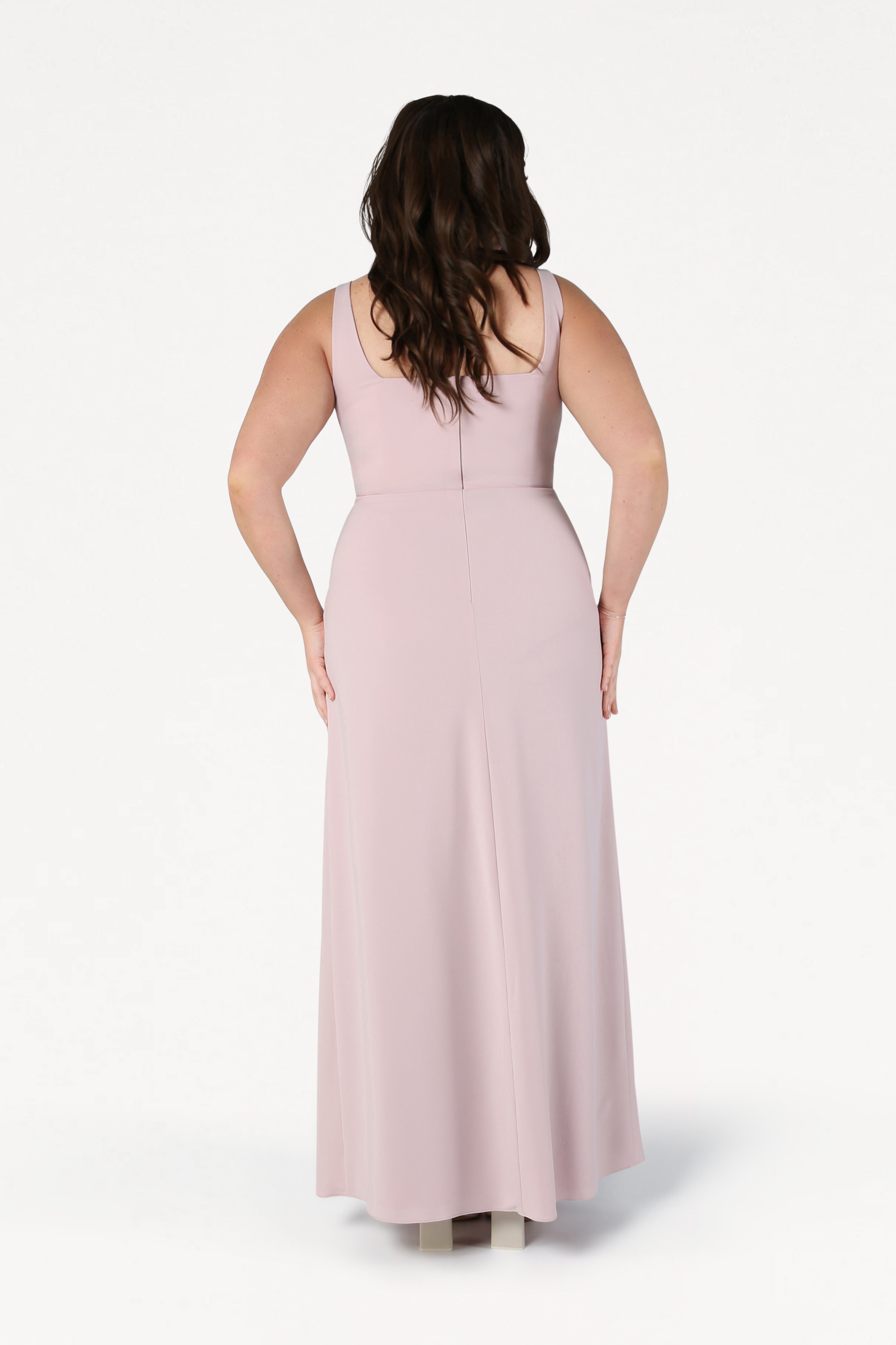 A woman with long brown hair stands with her back to the camera, wearing the TH&TH Helena Bridesmaid Dress in Smoked Blush and light-colored shoes against a white background.