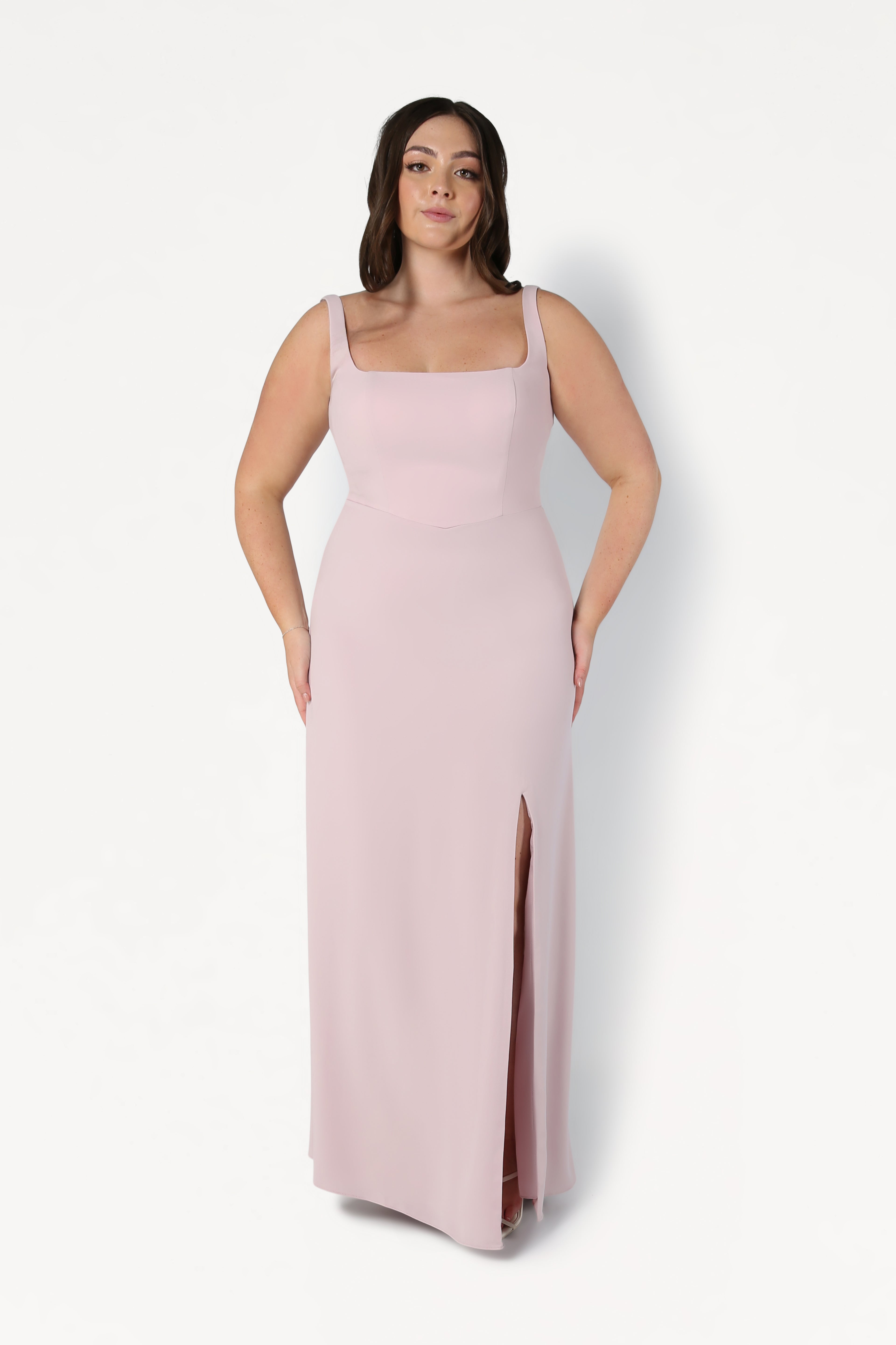 A woman wears the TH&TH Helena Bridesmaid Dress in Smoked Blush, a sleeveless, floor-length design with a square neckline and thigh-high slit, standing against a plain white background.