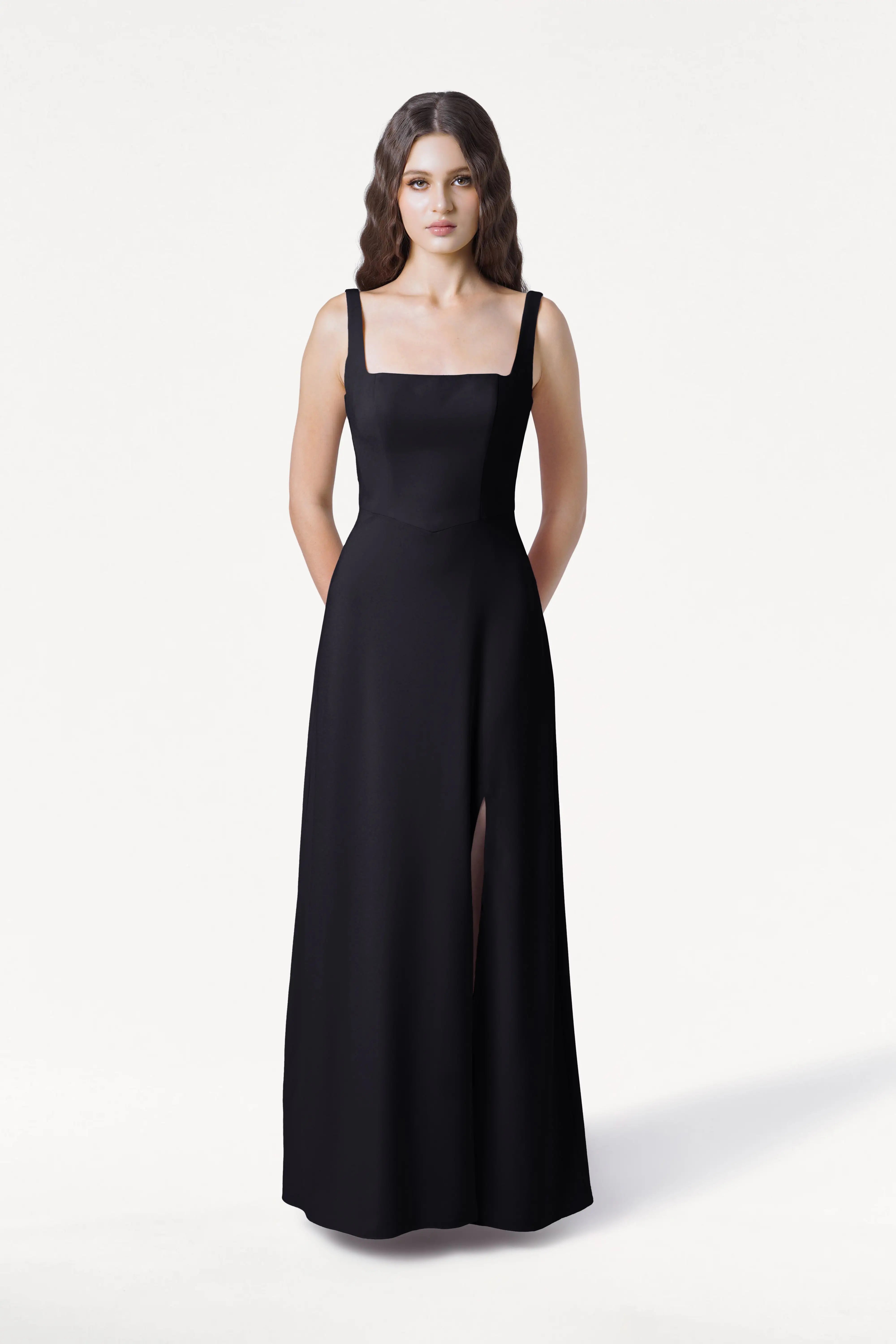 A woman with long wavy brown hair stands against a white background, wearing the TH&TH Helena Bridesmaid Dress in Black—a modern, size-inclusive, sleeveless gown with a square neckline and thigh-high slit. Her hands are behind her back.