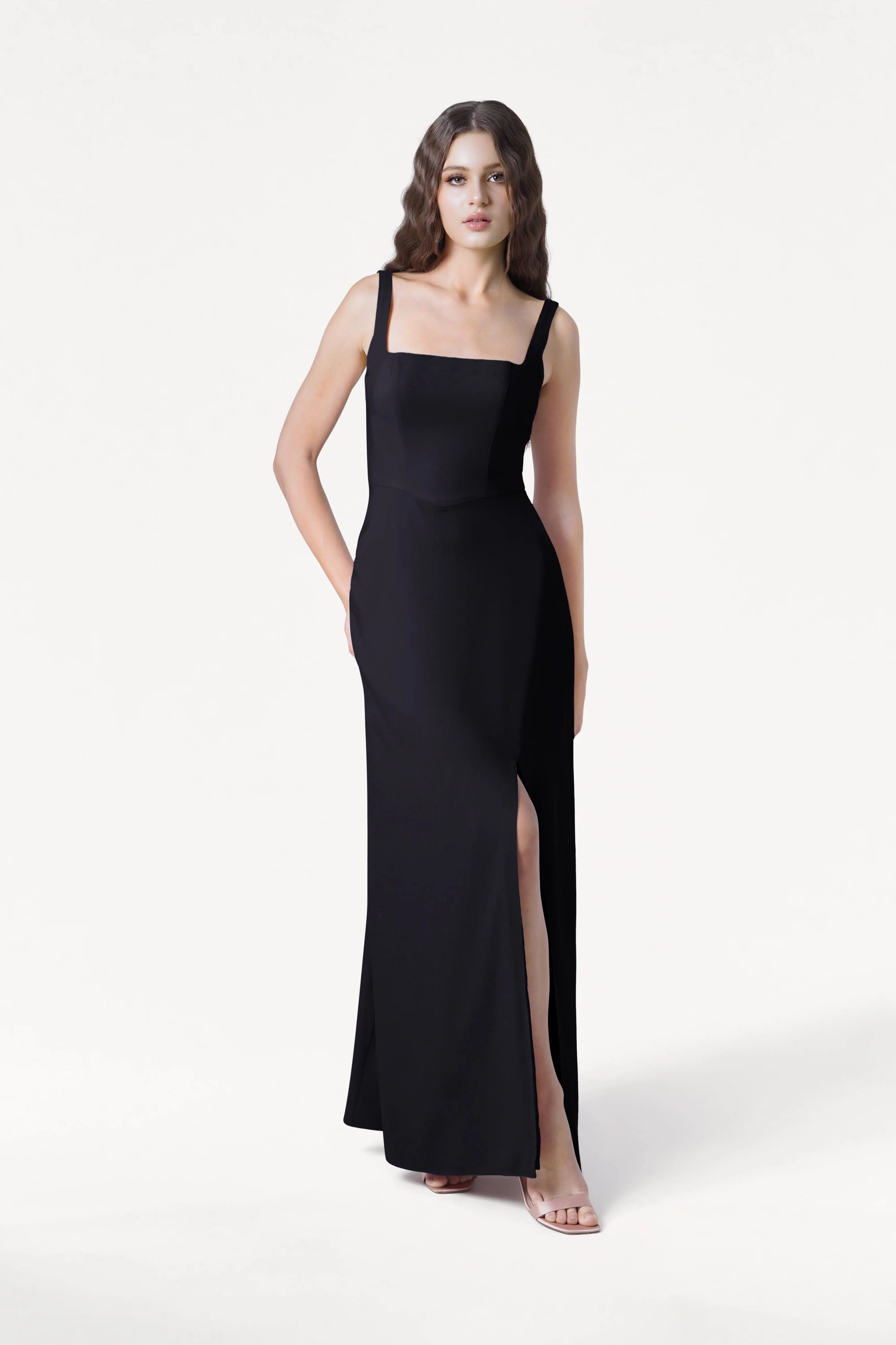A woman wears the Helena Bridesmaid Dress in Black by TH&TH—a sleeveless, floor-length moss crepe gown with a square neckline and high side slit. She pairs it with open-toe heels and poses against a plain white background.