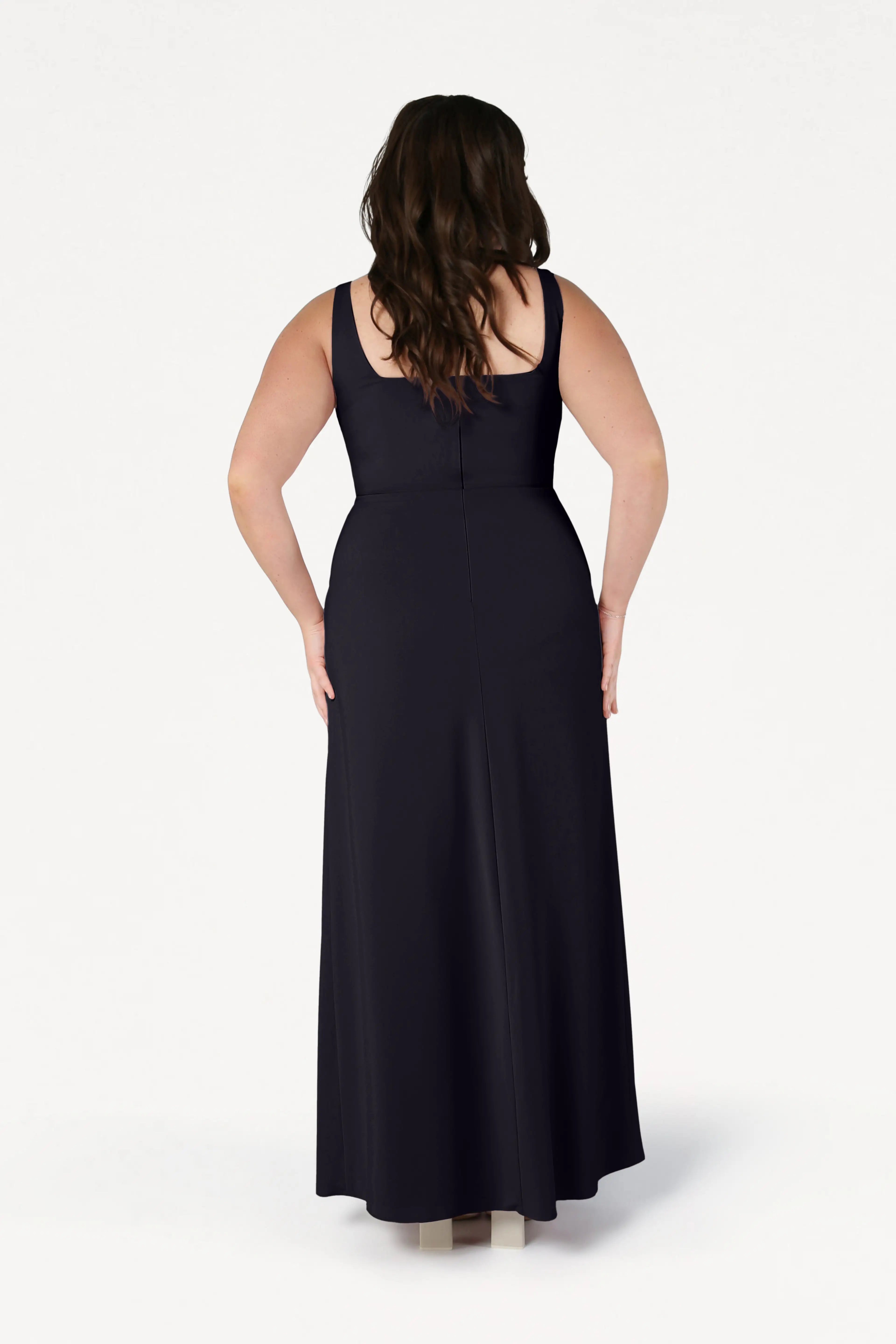 A woman with wavy brown hair, facing away, wears the sleeveless, floor-length Helena Bridesmaid Dress in Black by TH&TH and beige shoes against a plain white background.