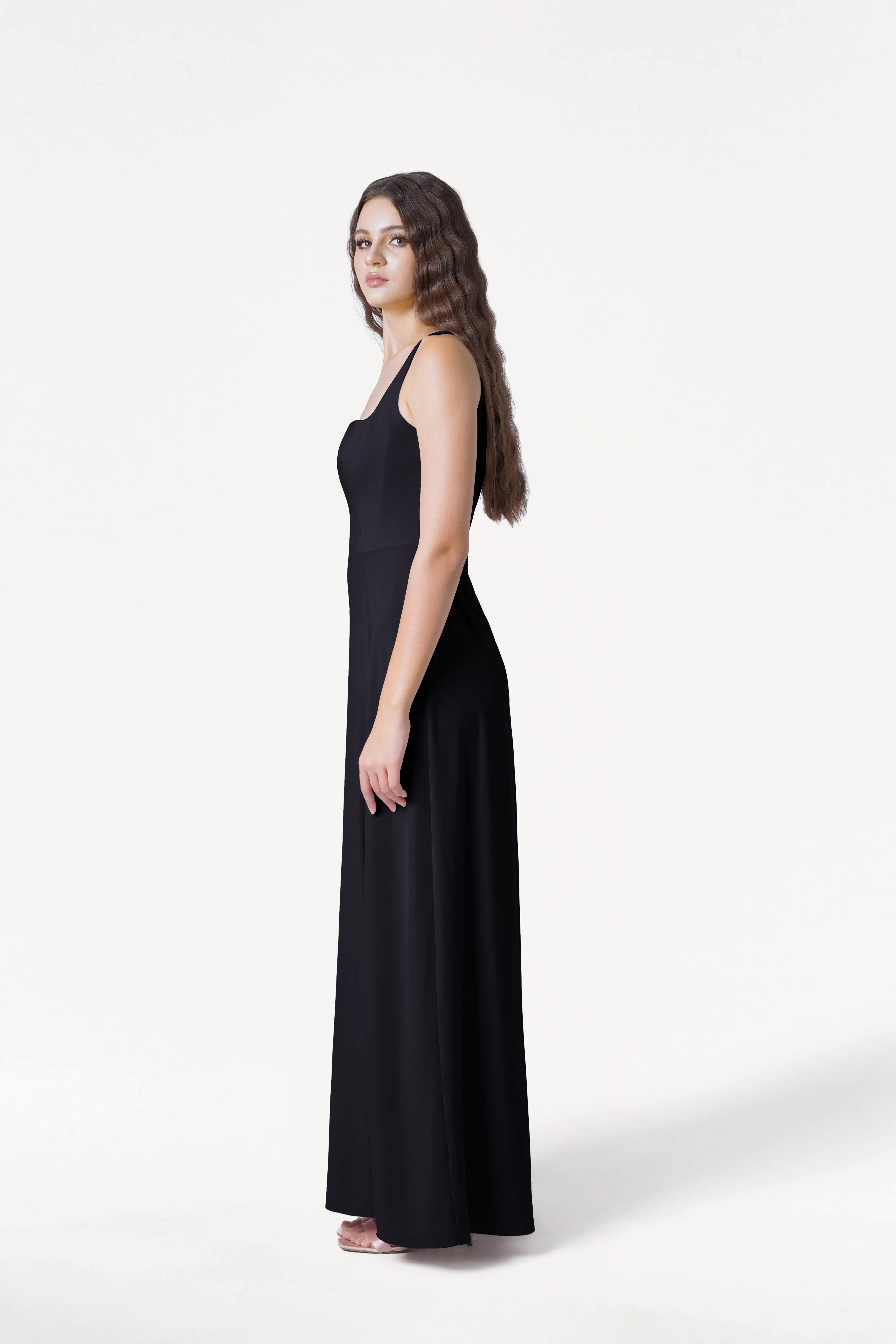 A woman with long wavy hair stands in profile, wearing the Helena Bridesmaid Dress in Black by TH&TH—a sleeveless gown paired with white sandals—set against a plain white background.