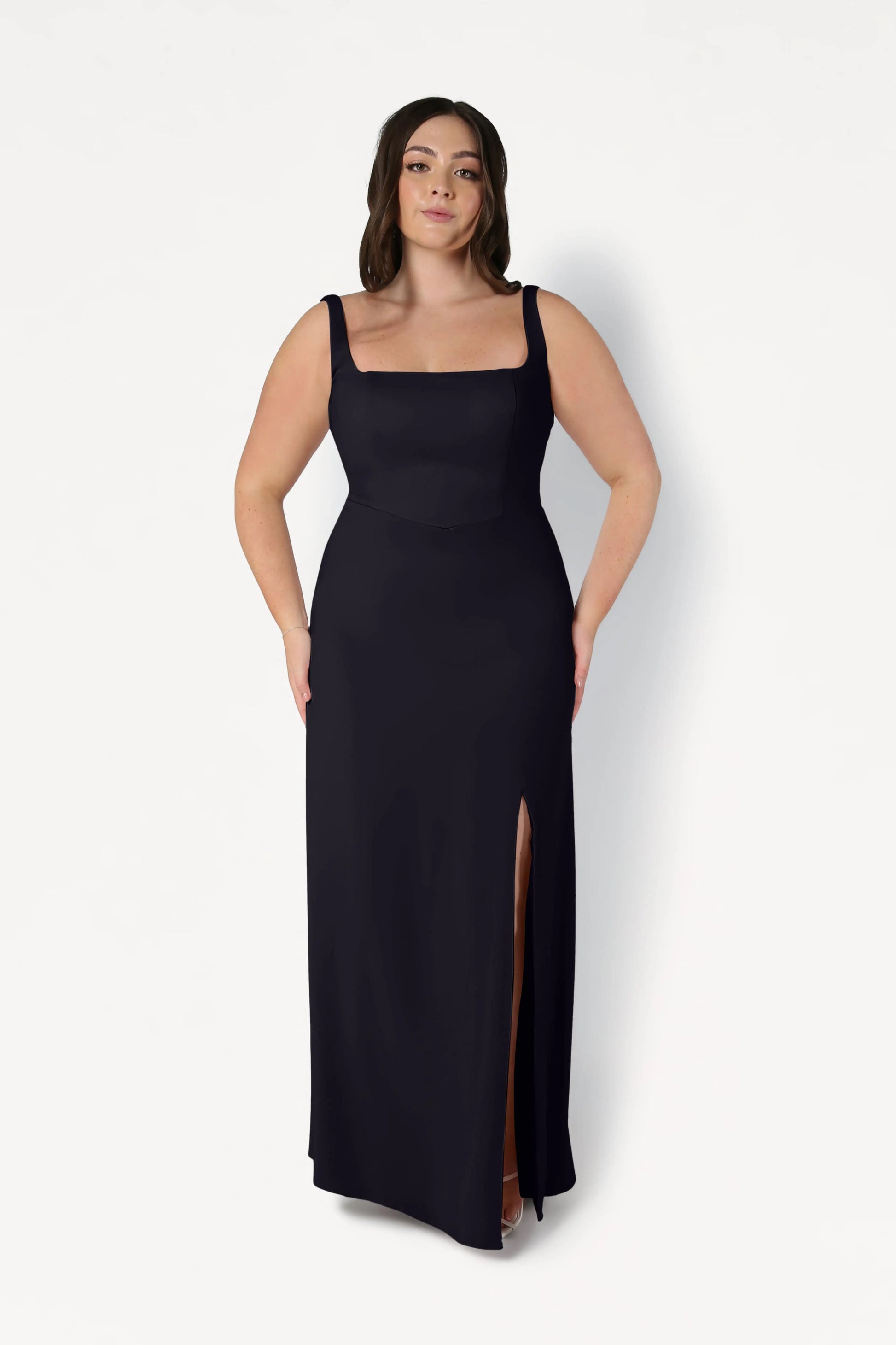 Woman with long brown hair wearing the TH&TH Helena Bridesmaid Dress in Black, a sleeveless, floor-length gown with a square neckline and high slit, standing against a plain white background.