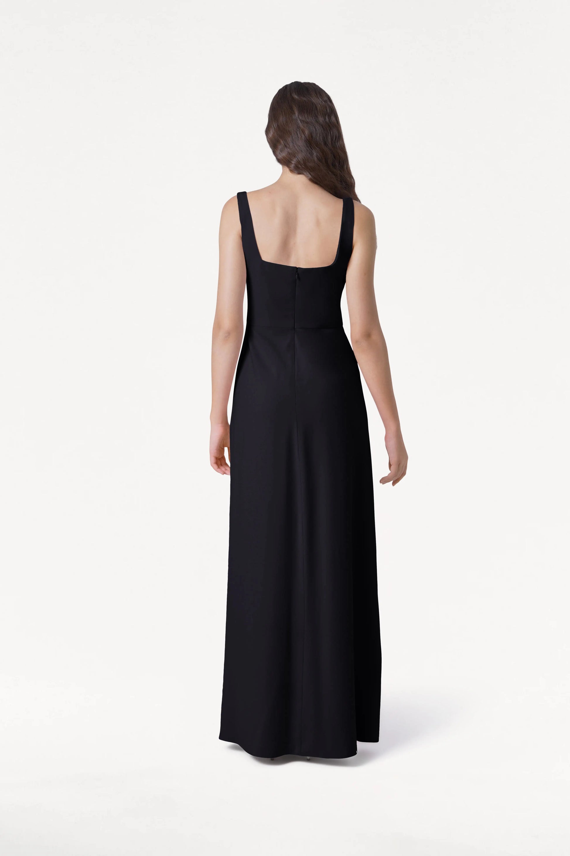 A woman with long brown hair, seen from the back, wears the floor-length Helena Bridesmaid Dress in Black by TH&TH against a plain white background.