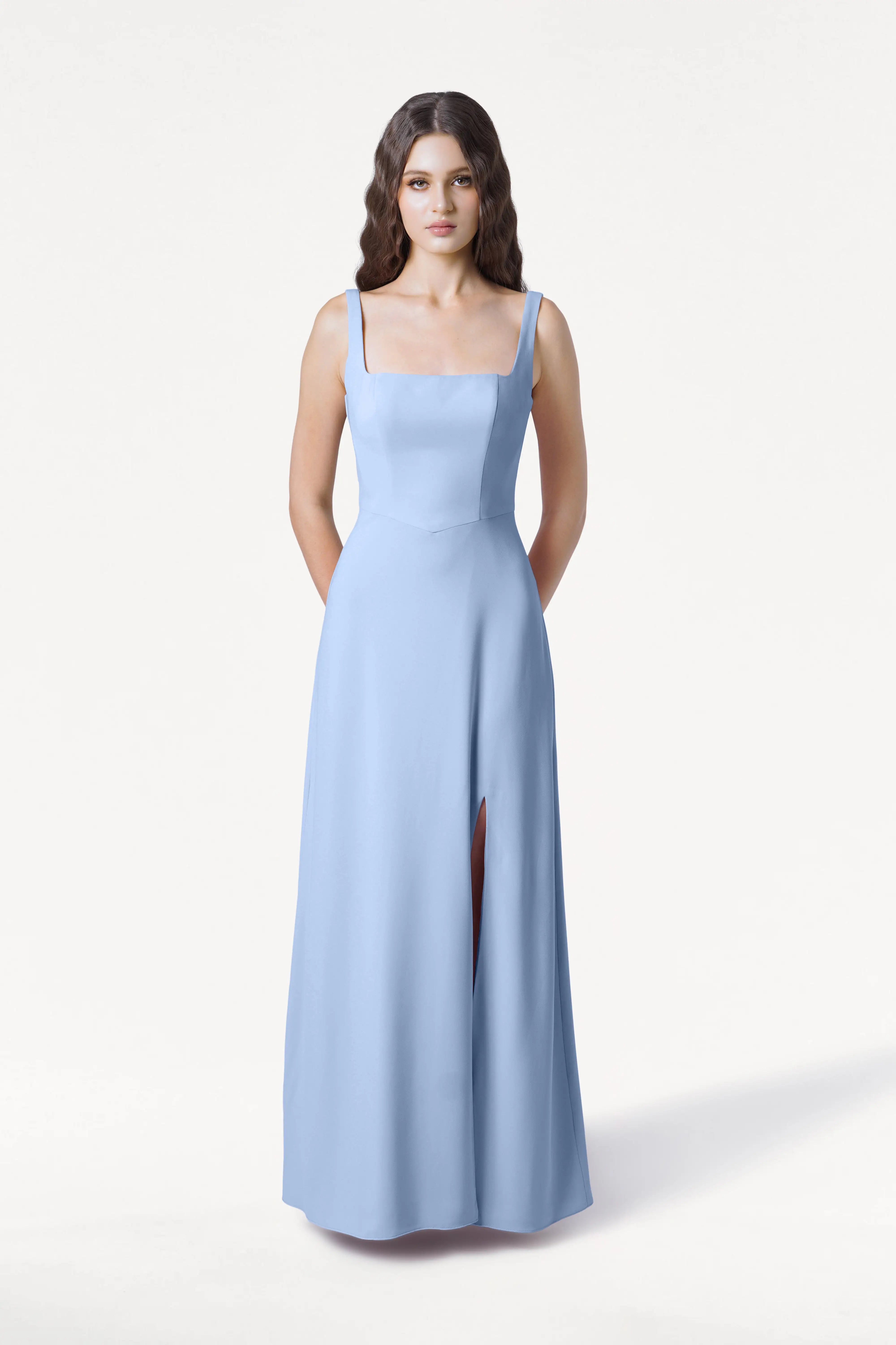 A woman faces forward wearing the Helena Bridesmaid Dress in Dusty Blue by TH&TH, a size-inclusive, floor-length moss crepe gown with a square neckline and high slit. Her hands are behind her back. She has long wavy brown hair. The background is plain white.