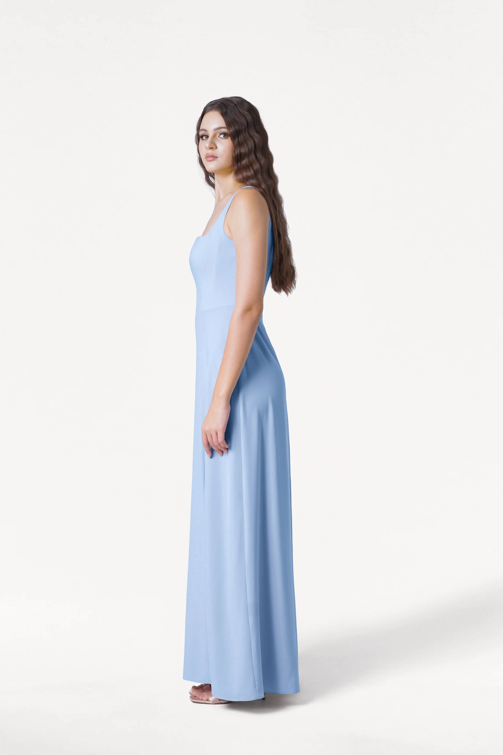 A woman with long, wavy brown hair stands sideways against a plain white background wearing the TH&TH Helena Bridesmaid Dress in Dusty Blue, a sleeveless, floor-length modern gown paired with white sandals.