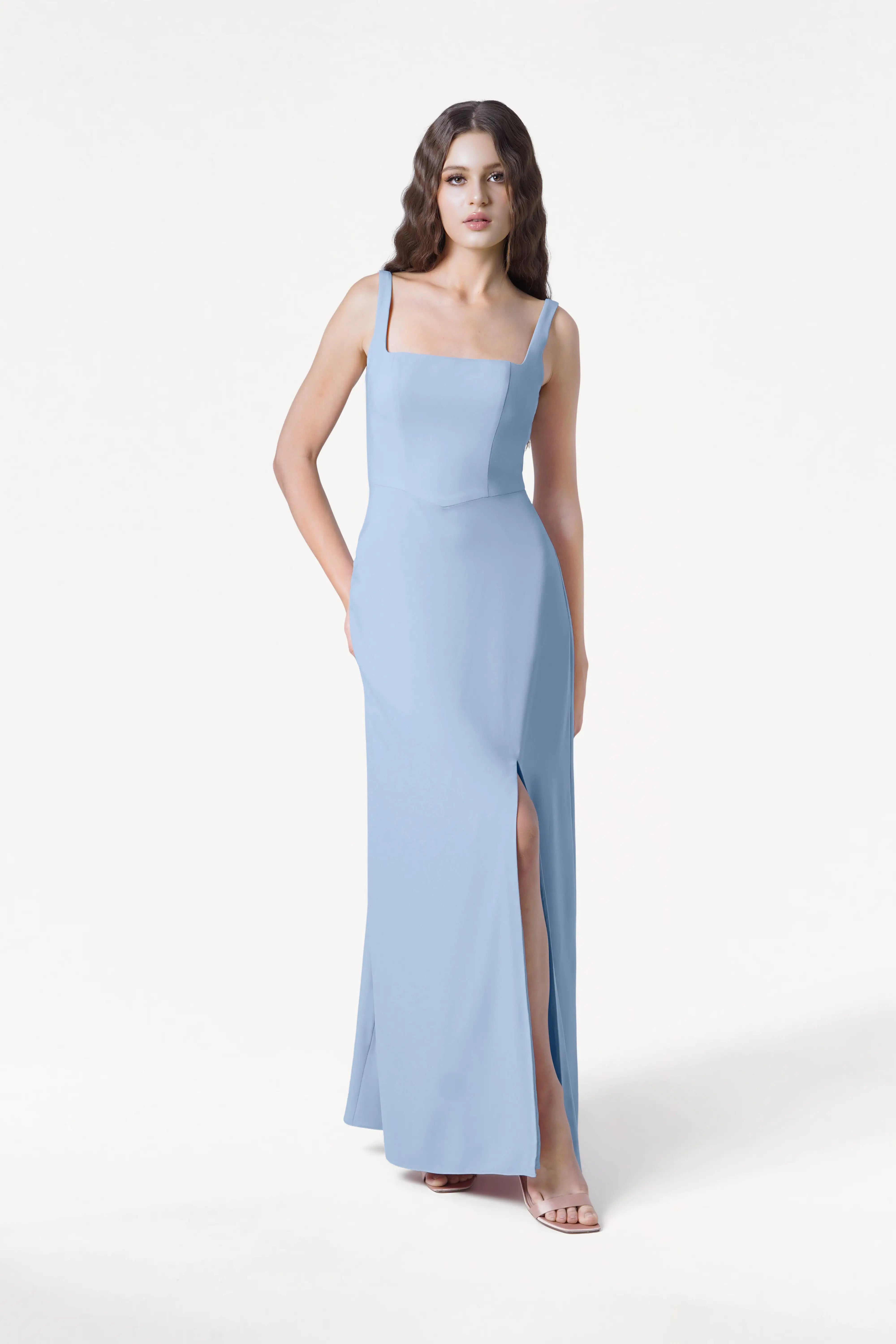 A woman with long brown hair wears the TH&TH Helena Bridesmaid Dress in Dusty Blue, a sleeveless, floor-length modern moss crepe gown with a square neckline and thigh-high slit. She stands against a plain white background in open-toed shoes.