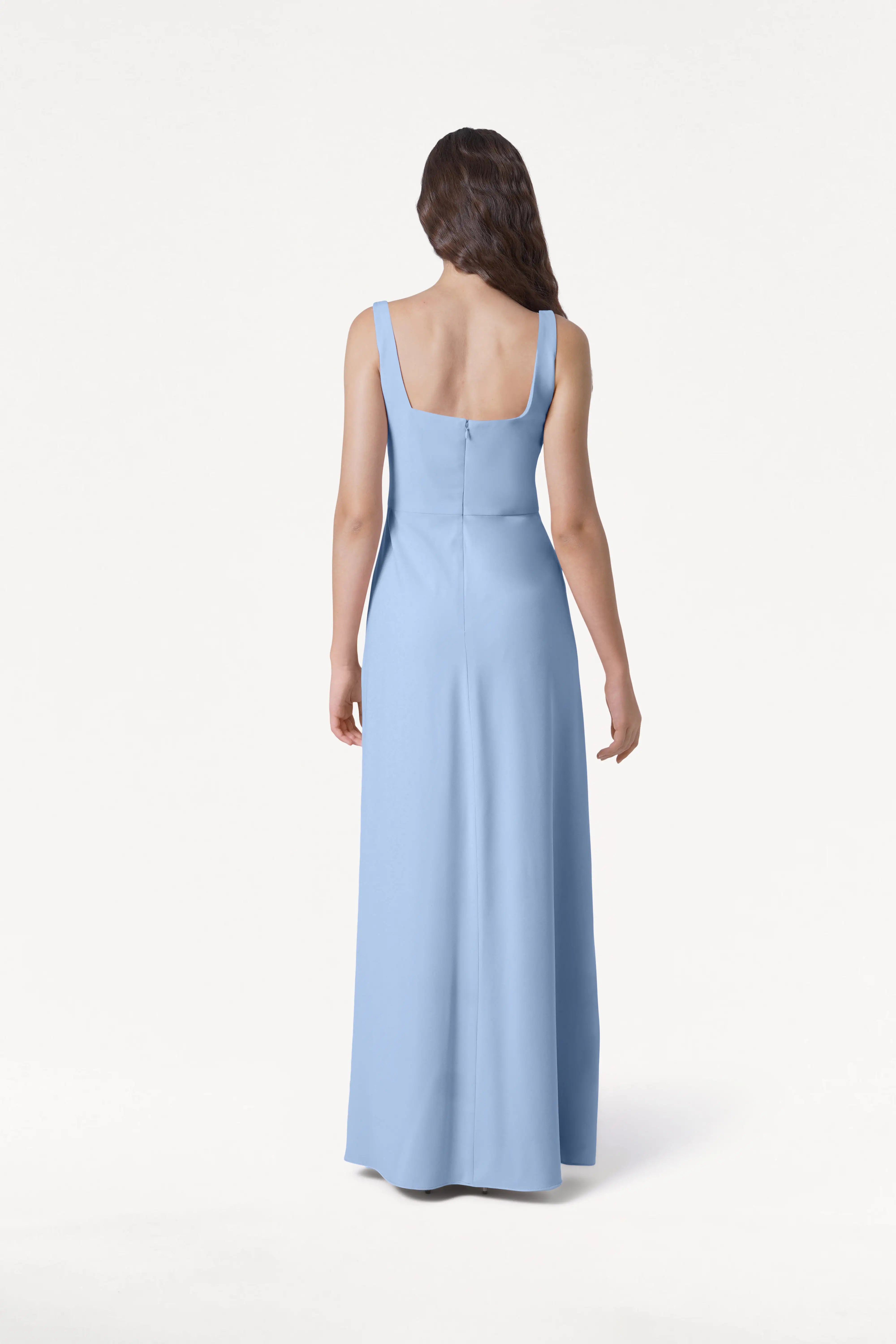 A woman with long wavy brown hair models the TH&TH Helena Bridesmaid Dress in Dusty Blue, featuring a sleeveless, floor-length design with a square neckline. She is shown from the back against a plain white background.