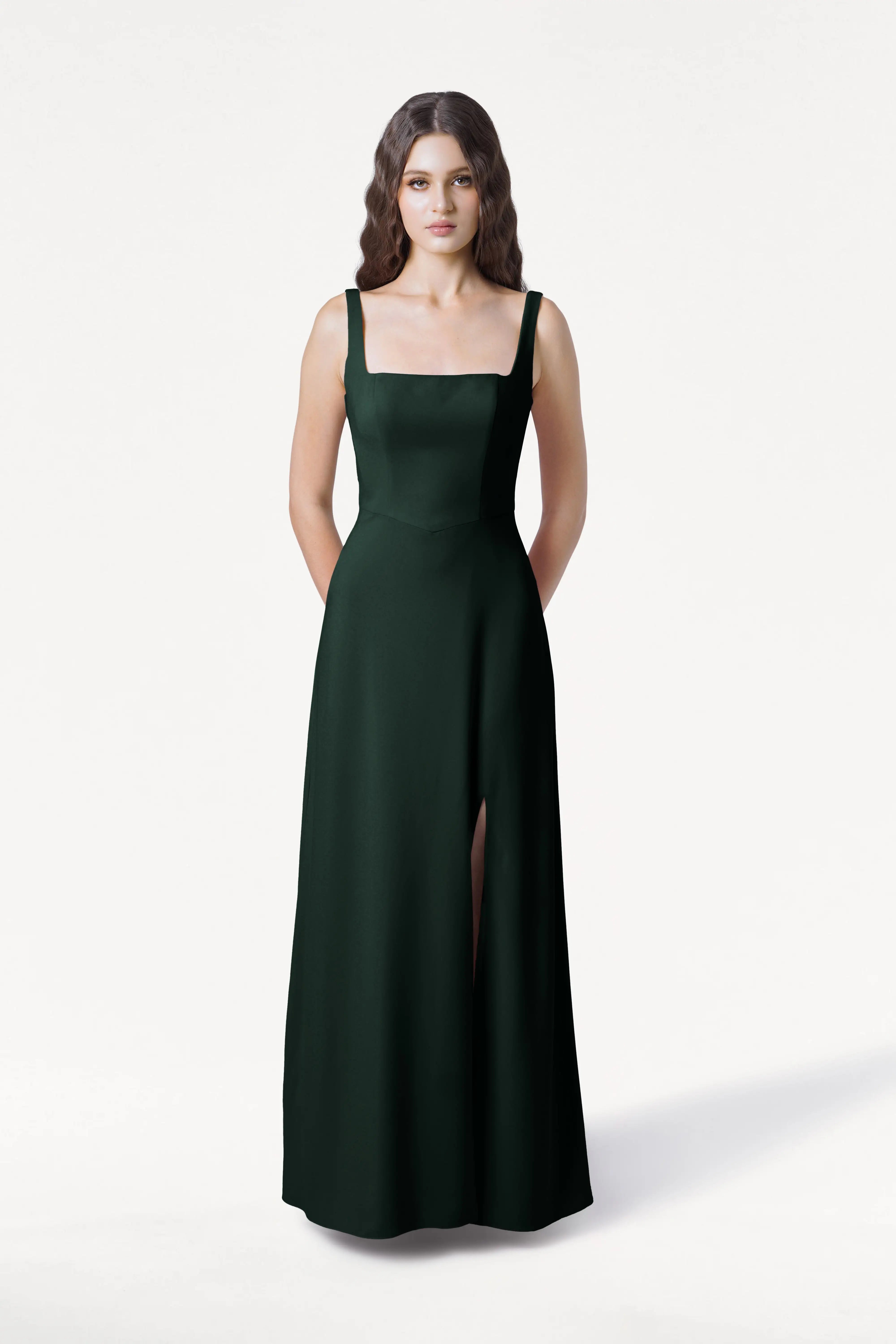 A woman with long, wavy brown hair stands facing forward in the size-inclusive TH&TH Helena Bridesmaid Dress in Forest Green, featuring a modern square neckline and thigh-high slit, against a plain white background.