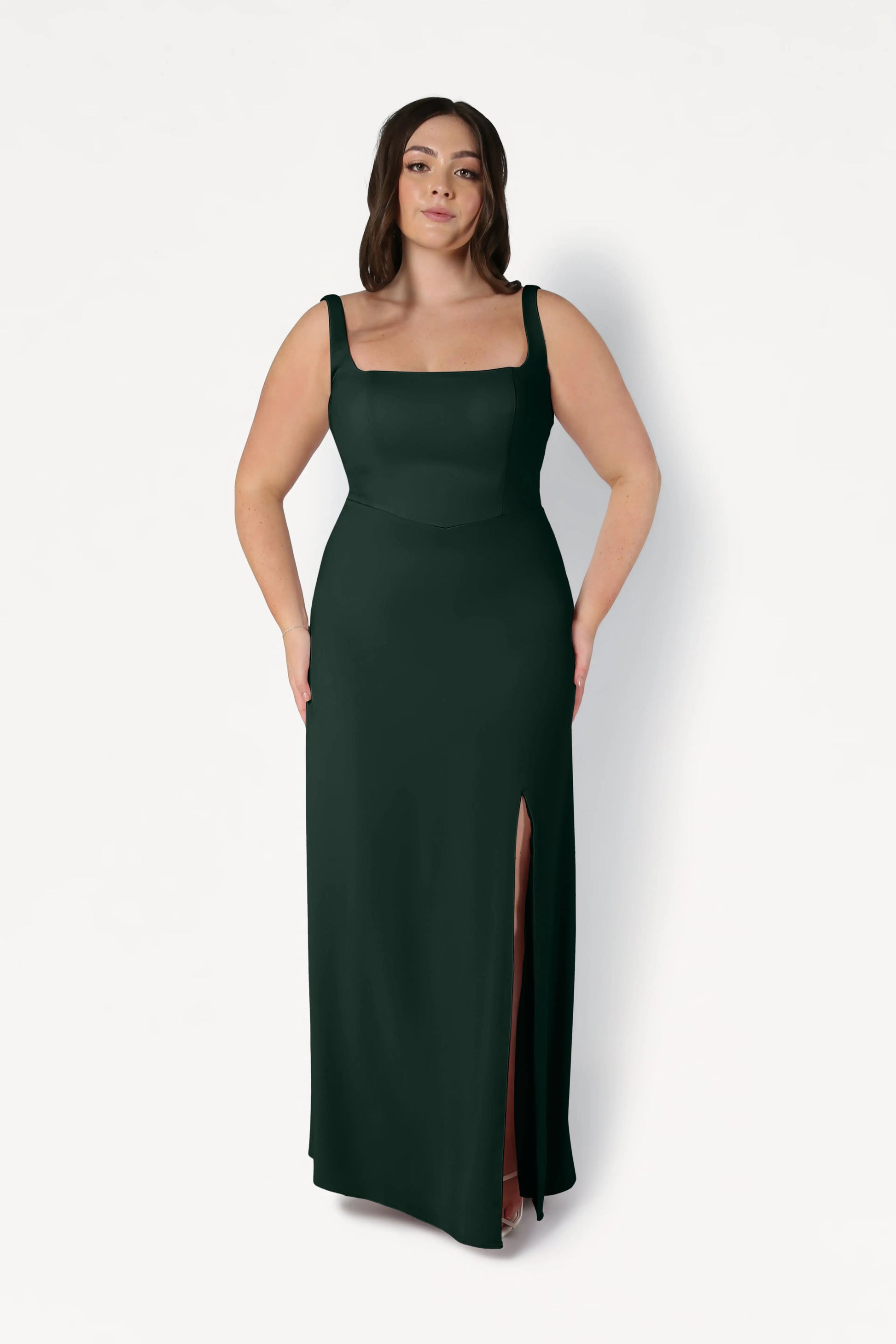 A woman with long brown hair models the TH&TH Helena Bridesmaid Dress in Forest Green—a size-inclusive modern crepe dress with a square neckline and thigh-high slit—standing against a plain white background.