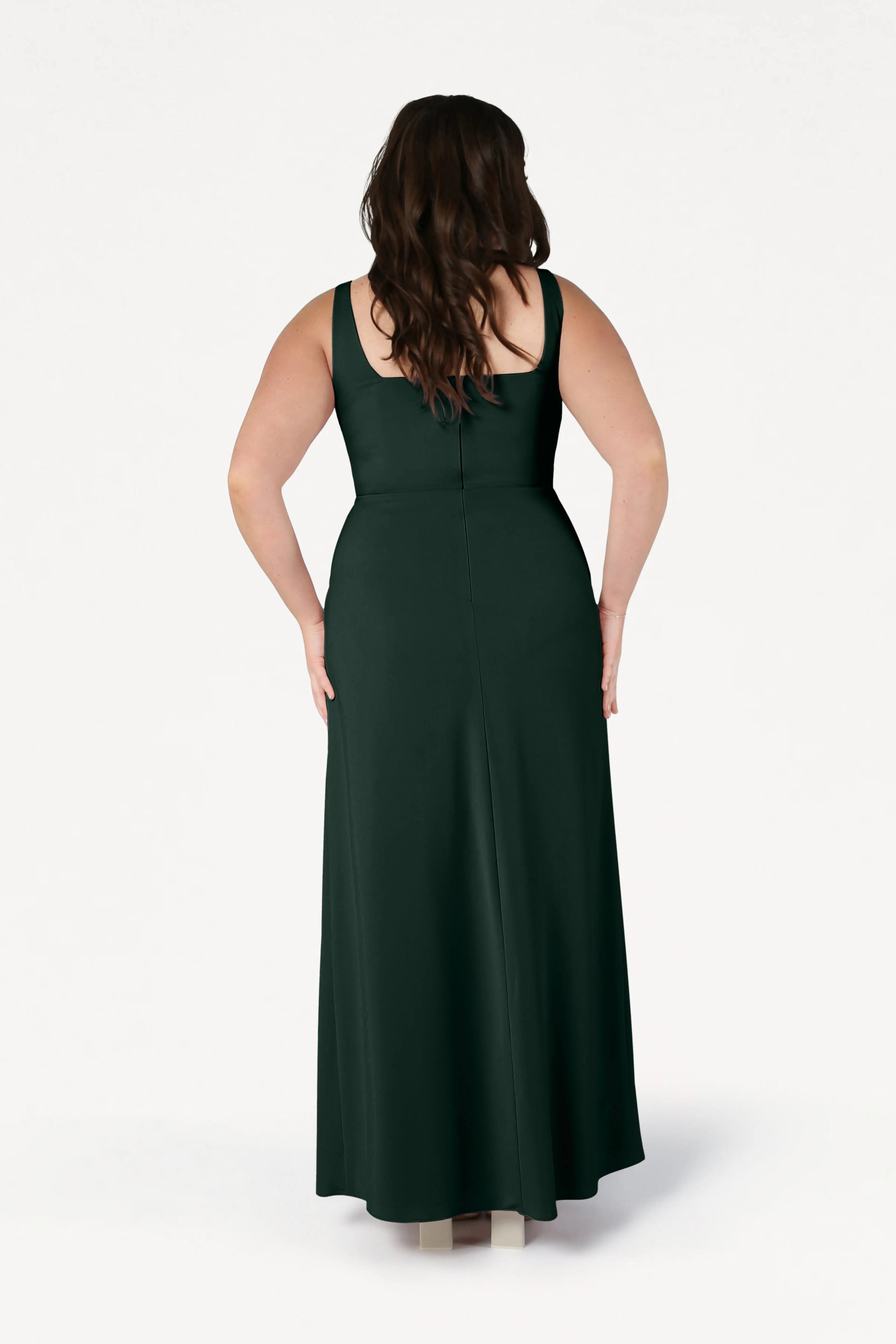 A woman with long brown hair stands with her back to the camera against a white background, wearing the sleeveless, floor-length Helena Bridesmaid Dress in Forest Green by TH&TH.