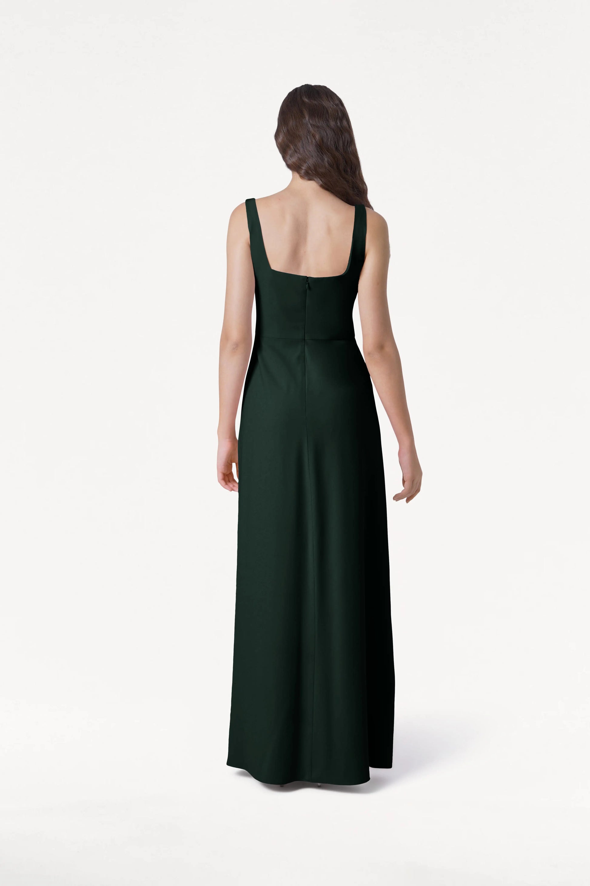 A woman with long wavy hair, seen from behind, wears the TH&TH Helena Bridesmaid Dress in Forest Green—sleeveless and floor-length with a square neckline and fitted bodice—standing against a plain white background.