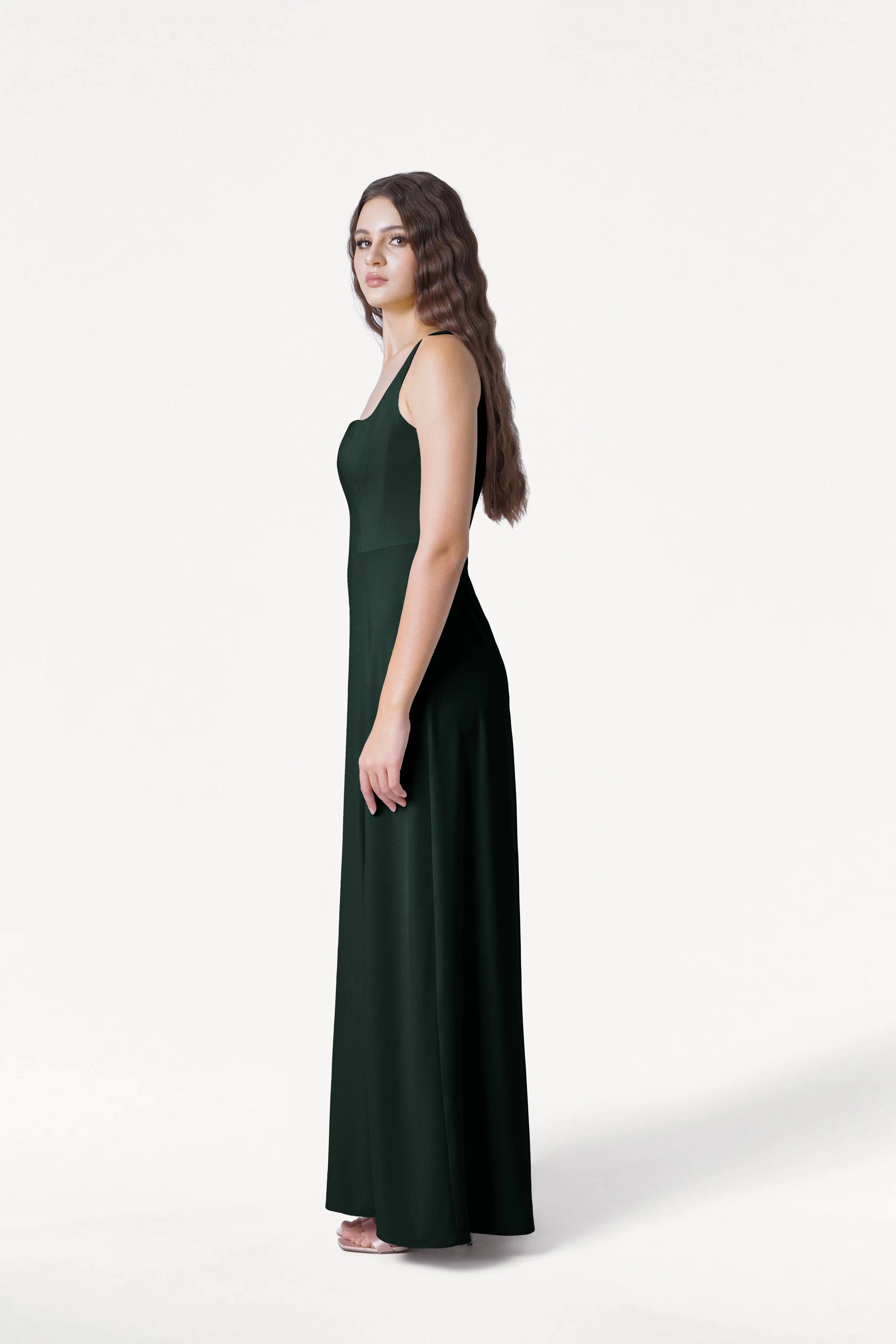 A young woman with long wavy hair stands in profile wearing the TH&TH Helena Bridesmaid Dress in Forest Green and white shoes against a plain white background.