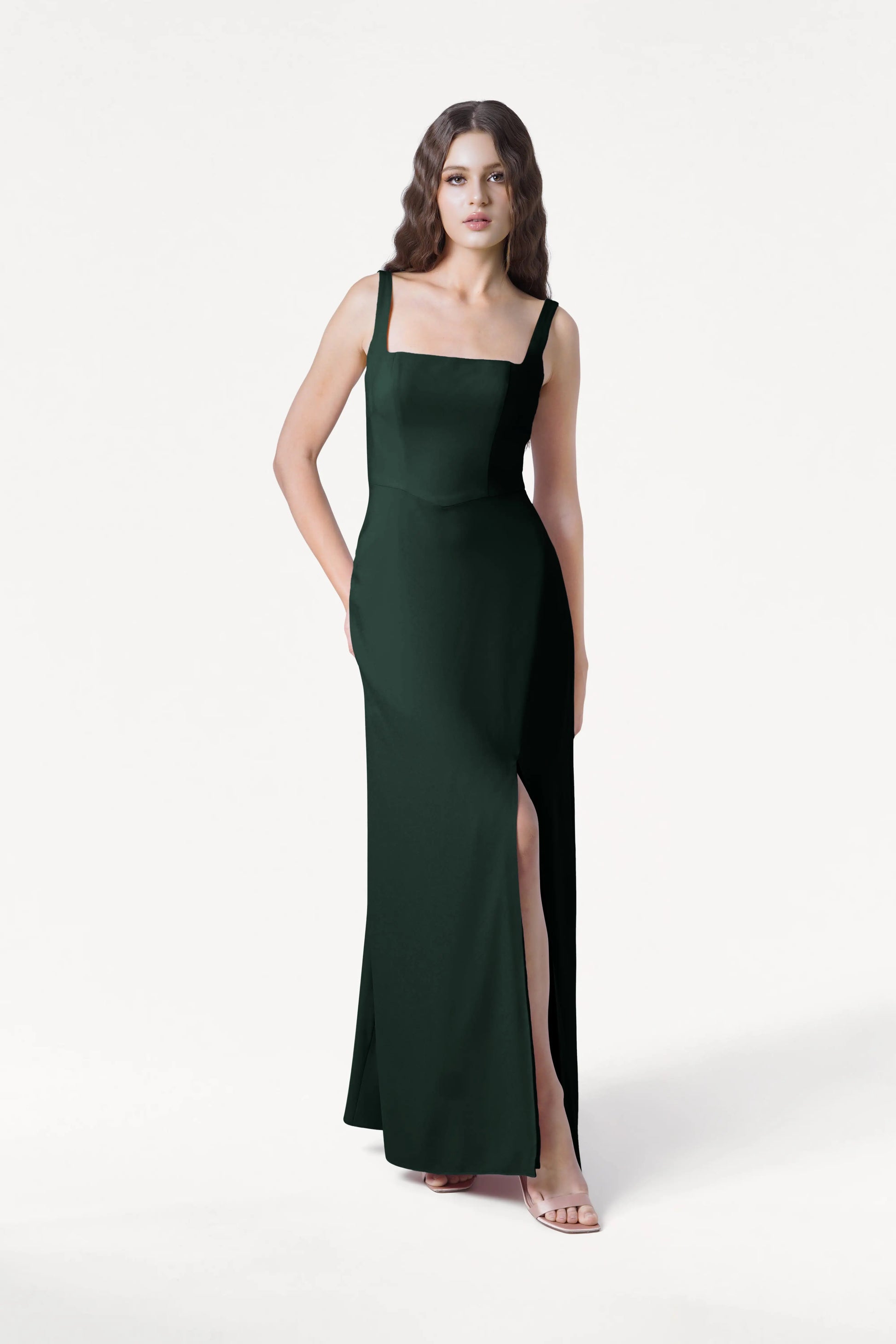 A woman stands against a white background in TH&TH’s Helena Bridesmaid Dress in Forest Green, featuring a square neckline, high slit, and sleeveless moss crepe design—an elegant pick for modern bridesmaids.