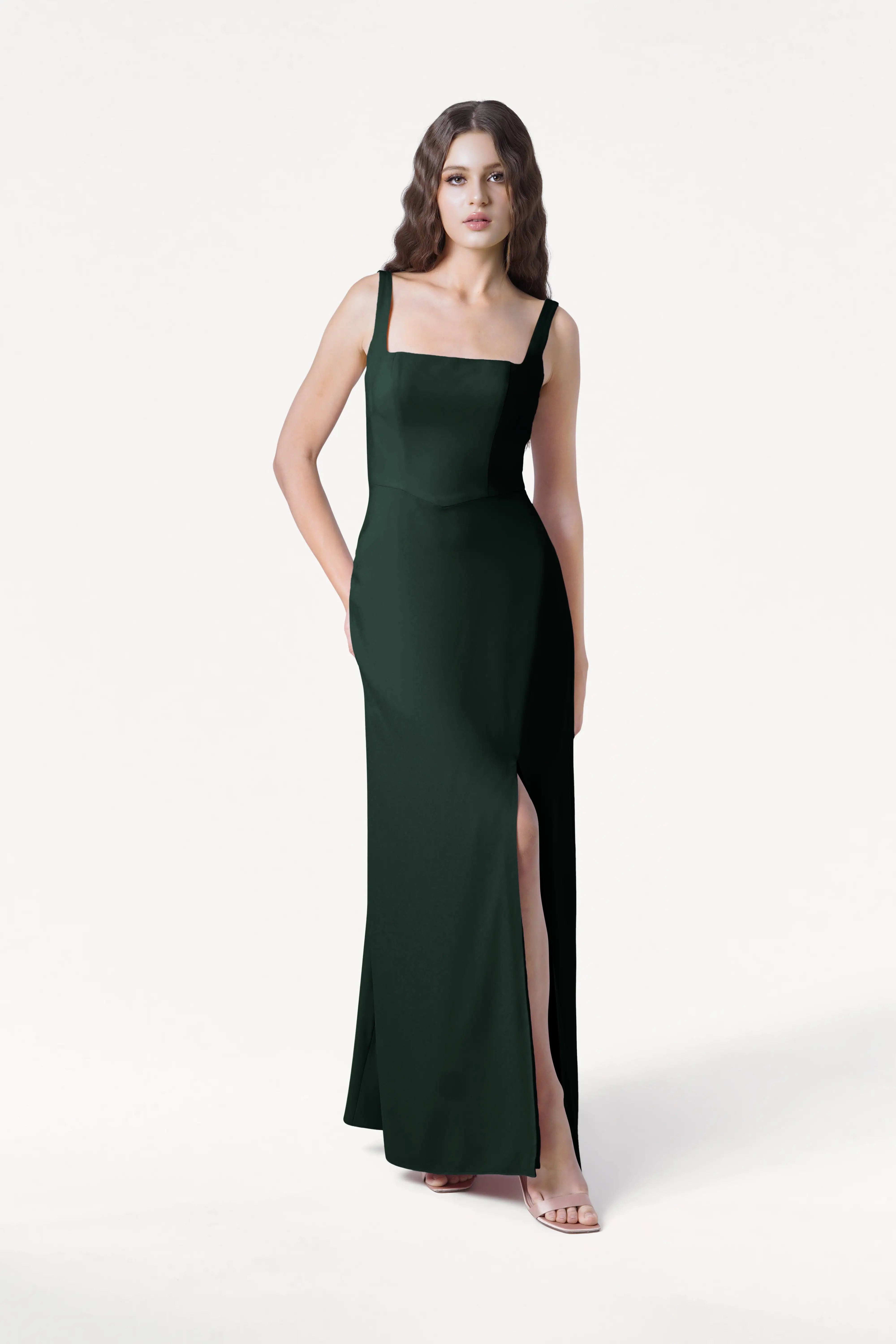 A woman stands against a white background in TH&TH’s Helena Bridesmaid Dress in Forest Green, featuring a square neckline, high slit, and sleeveless moss crepe design—an elegant pick for modern bridesmaids.