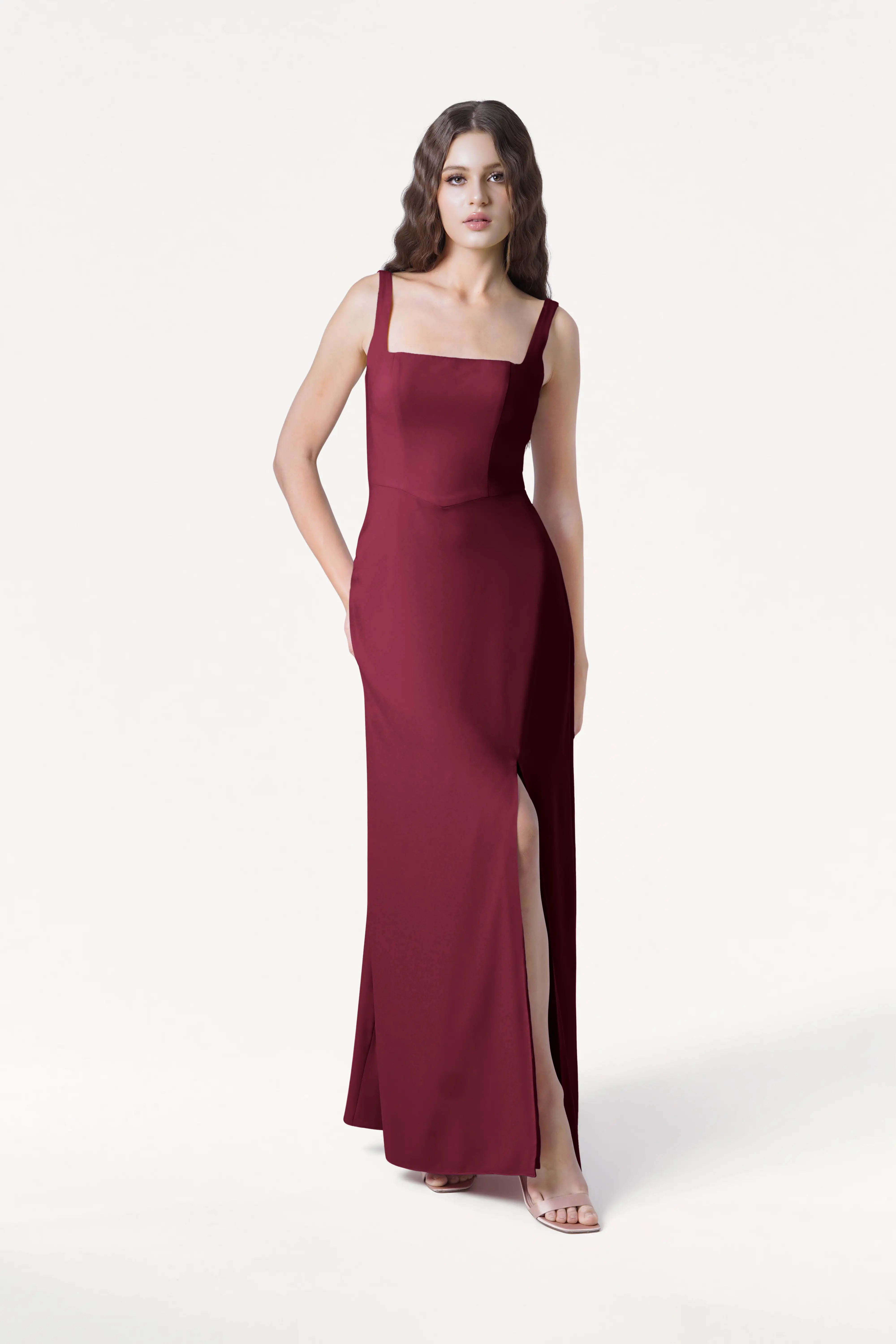 A woman with wavy brown hair models the size-inclusive TH&TH Helena Bridesmaid Dress in Merlot, featuring a square neckline, high slit, and floor-length design. She stands against a white background with one hand by her side and one behind her.