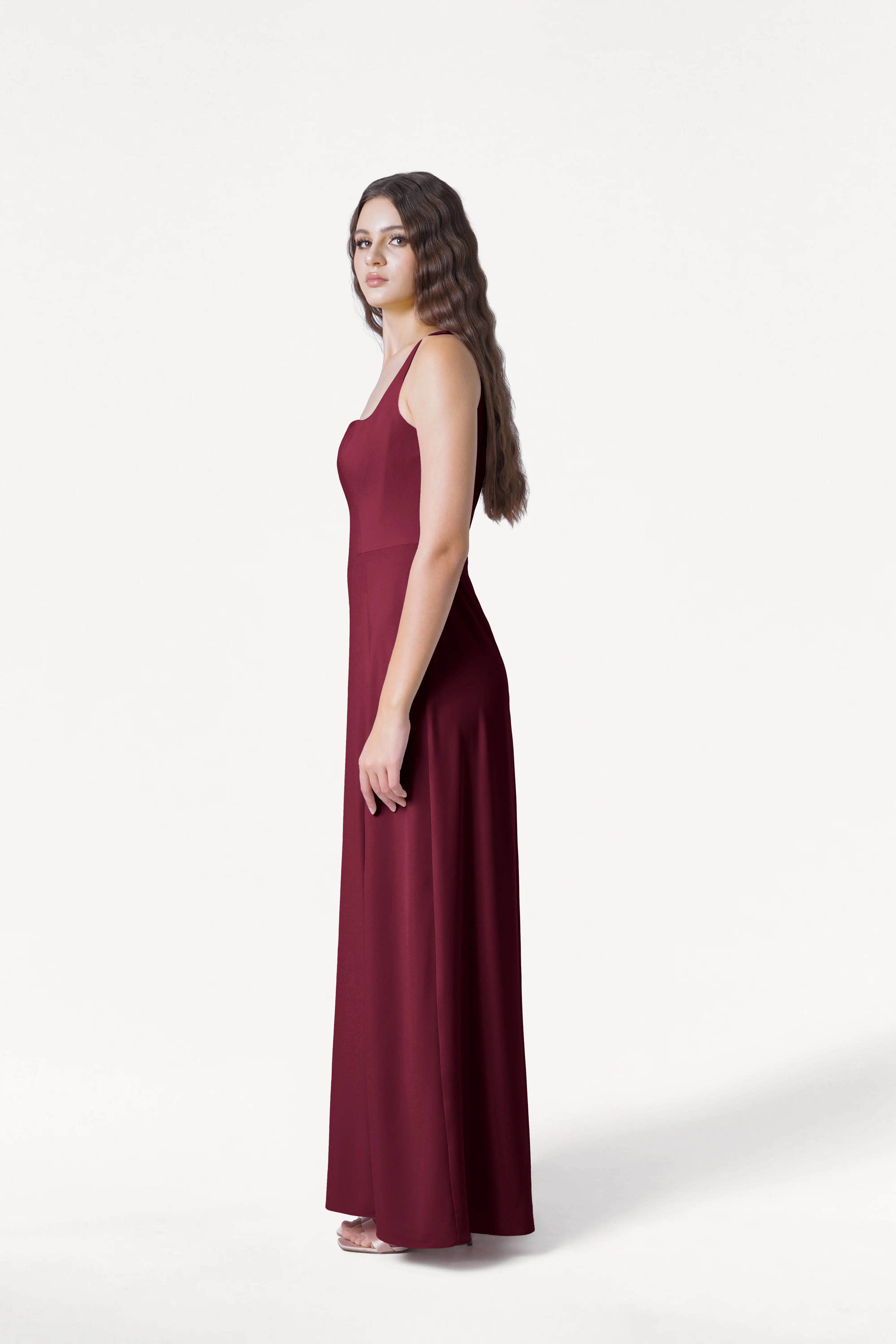 A woman with long wavy brown hair stands in profile, wearing the TH&TH Helena Bridesmaid Dress in Merlot—a modern, size-inclusive, floor-length gown with thin straps—set against a plain white background.