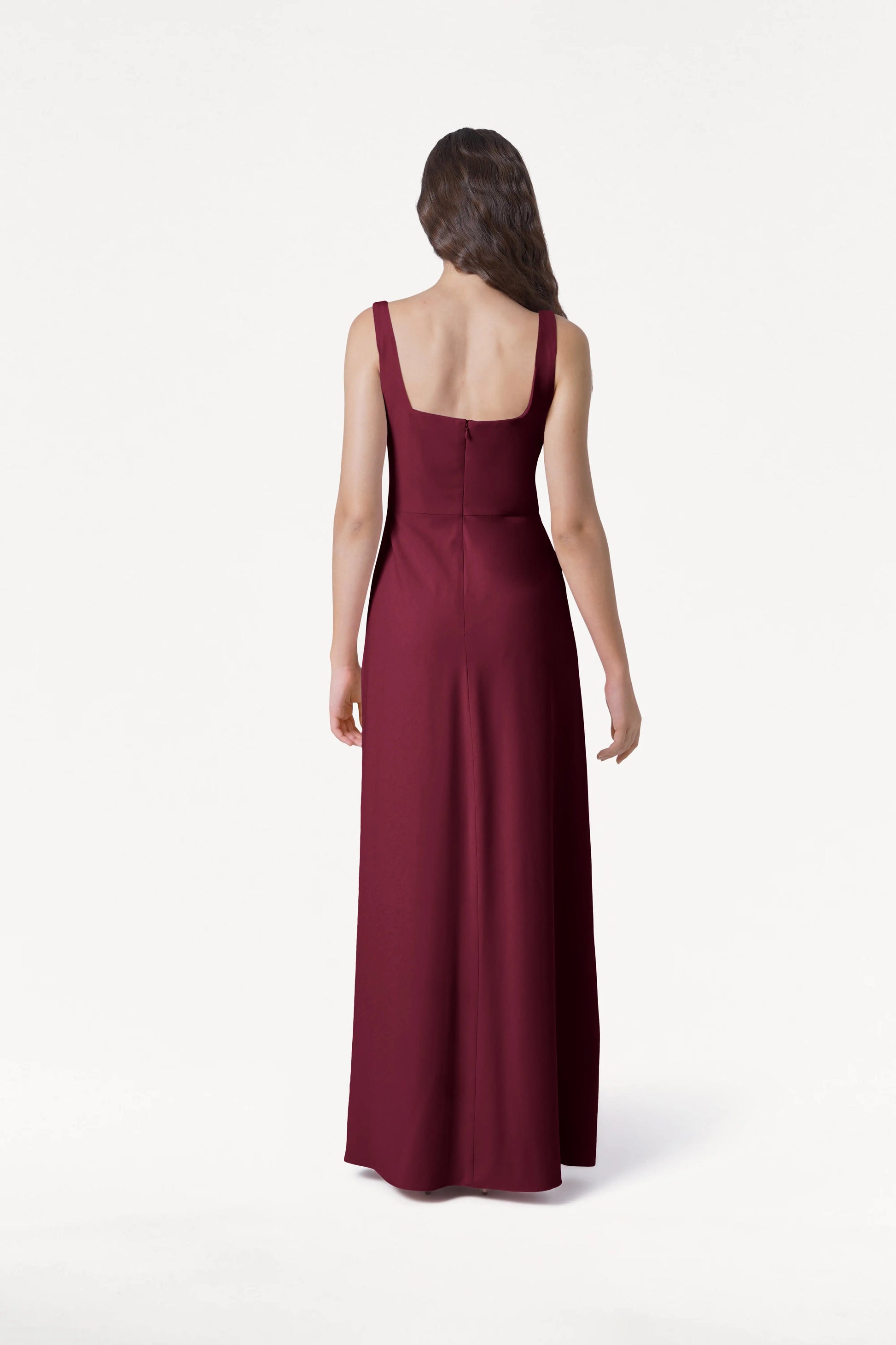 A woman with long, wavy brown hair faces away, wearing the TH&TH Helena Bridesmaid Dress in Merlot—sleeveless, floor-length with a square neckline and fitted bodice—against a plain white background.