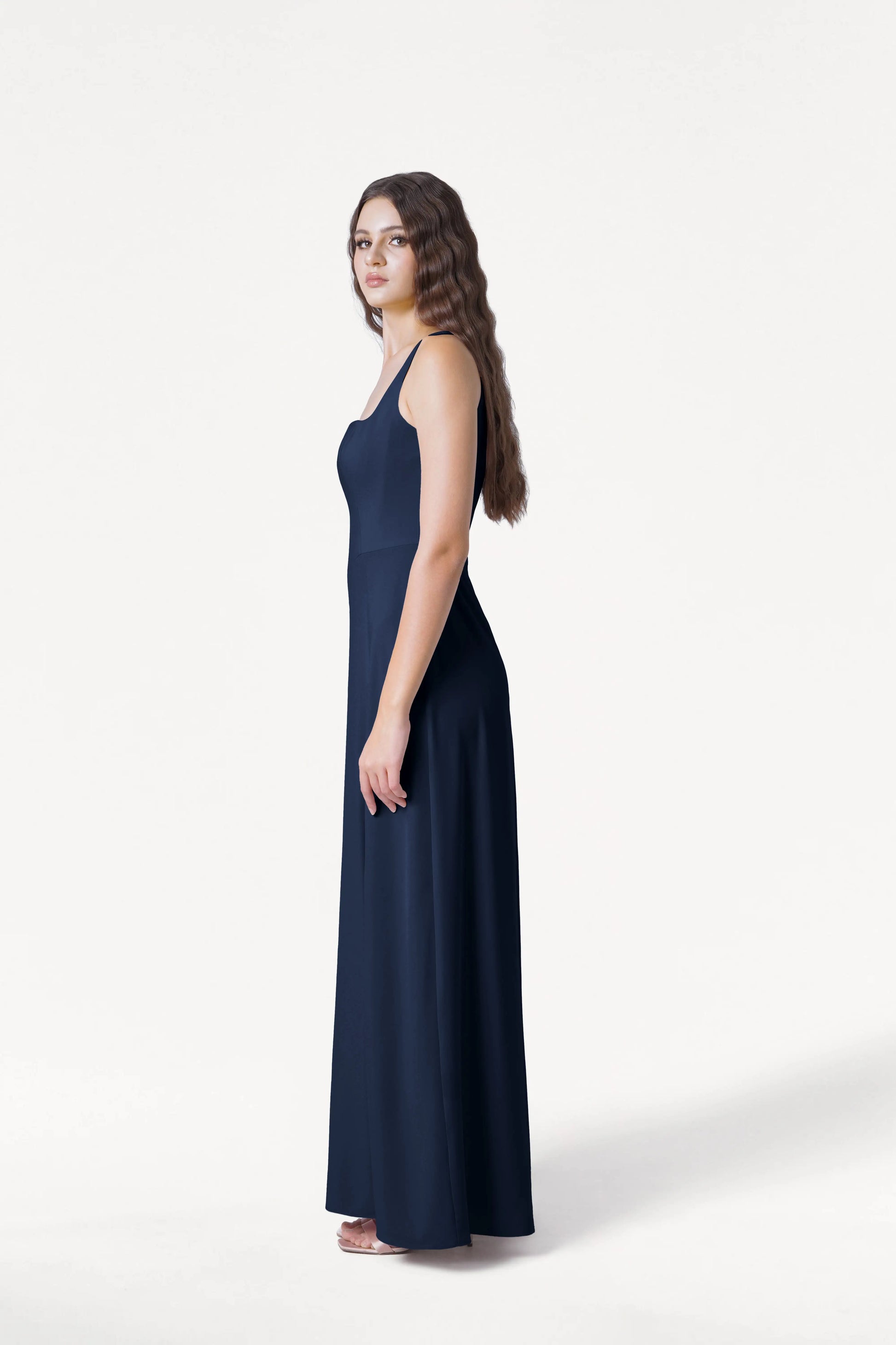 A woman with long, wavy brown hair stands in profile against a white background, wearing the TH&TH Helena Bridesmaid Dress in Navy—a size-inclusive, sleeveless, floor-length gown—styled with white heeled sandals.