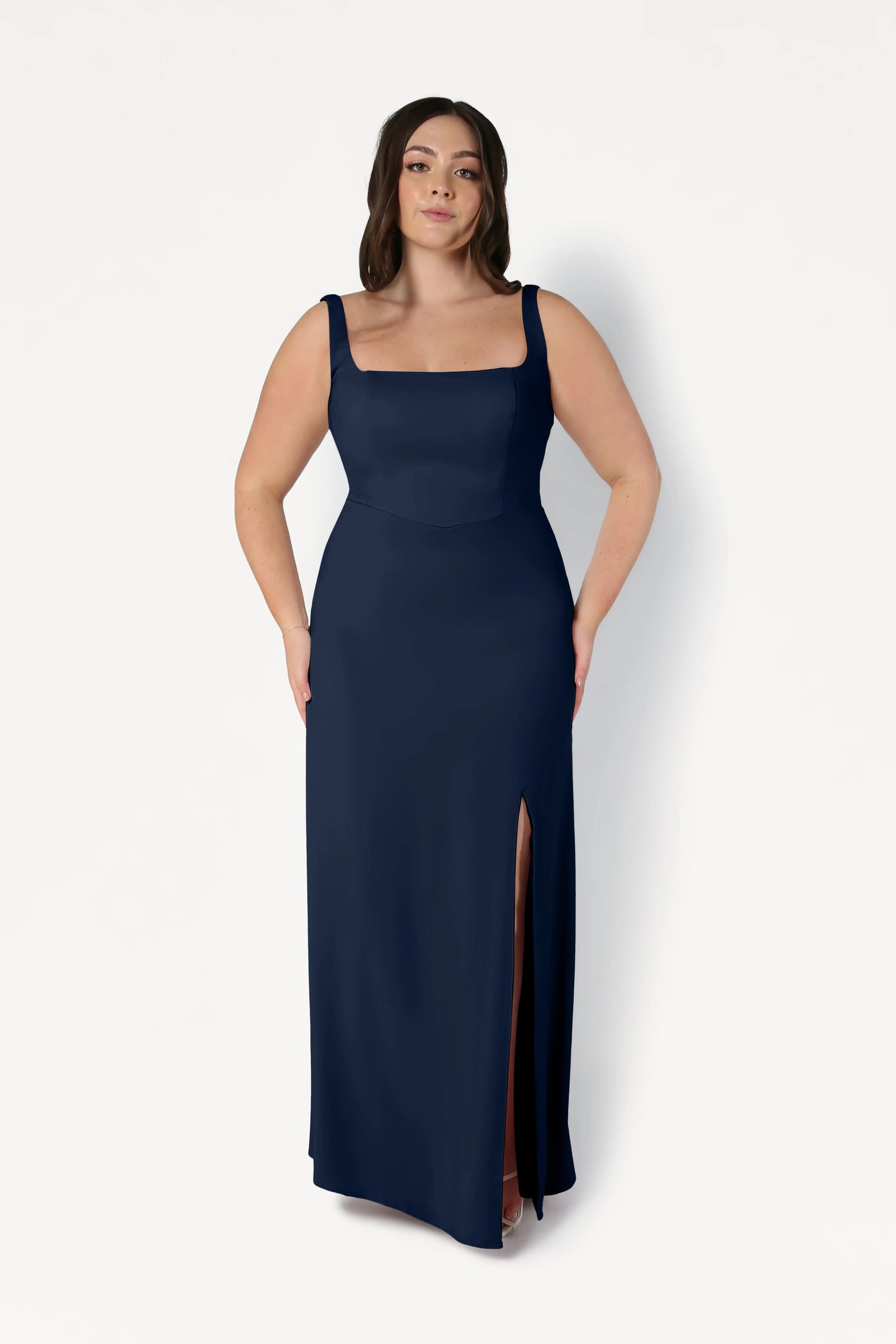 A woman wears the Helena Bridesmaid Dress in Navy by TH&TH, a sleeveless moss crepe gown with a square neckline and thigh-high slit, standing against a plain white background.