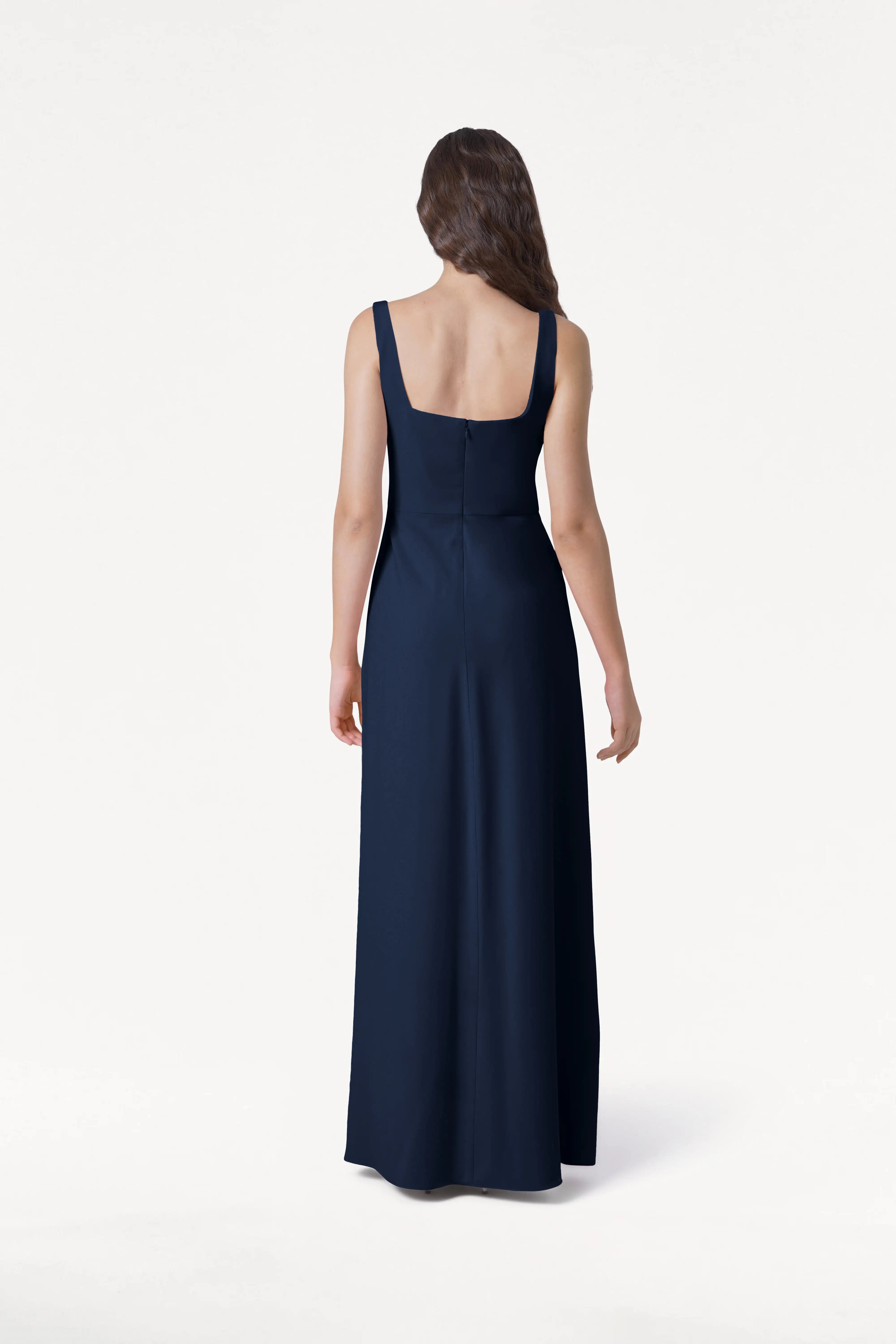 A woman with long brown hair faces away, wearing the TH&TH Helena Bridesmaid Dress in Navy: a sleeveless, floor-length gown with a square back neckline, set against a plain white background.