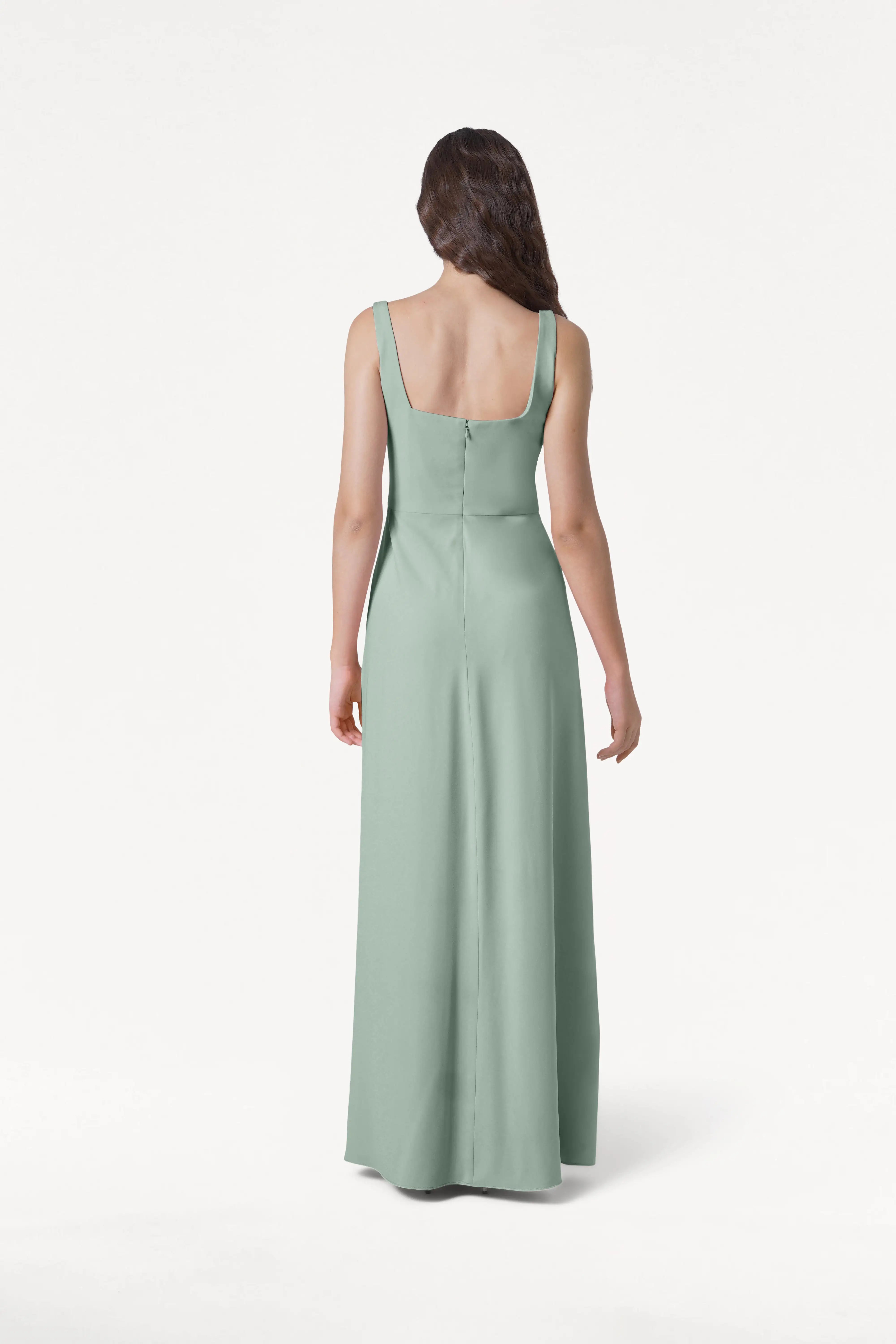 A woman with long, wavy brown hair stands facing away, wearing the TH&TH Helena Bridesmaid Dress in Sage Green, featuring a modern floor-length silhouette, square back, and center seam, set against a plain white background.
