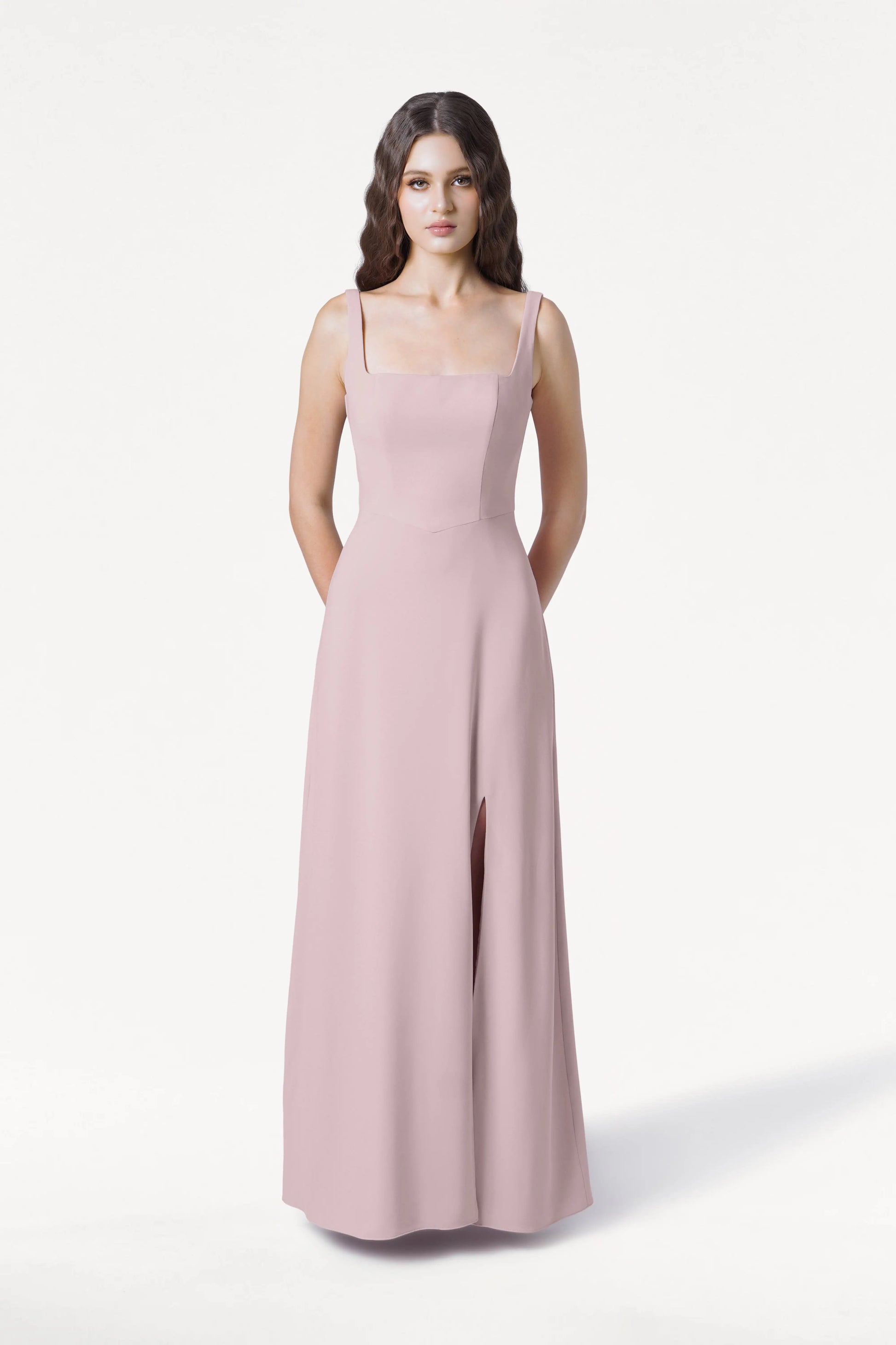 A woman with long brown hair wears the TH&TH Helena Bridesmaid Dress in Smoked Blush—a sleeveless, floor-length gown with a square neckline and front slit—standing facing forward against a plain white background.