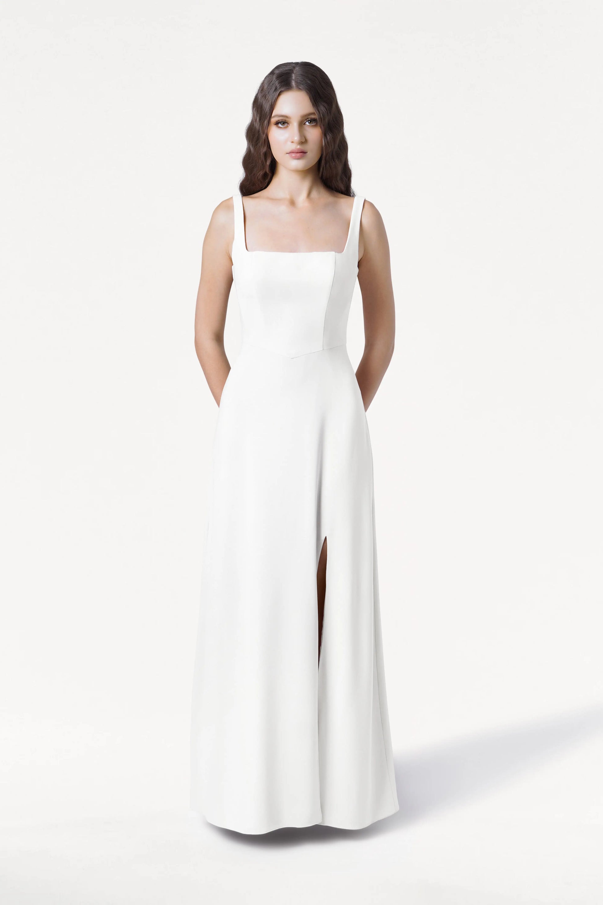 A woman with long, wavy dark hair stands against a white background, wearing the TH&TH Helena Dress in Ivory—a modern, sleeveless bridesmaid gown featuring a square neckline and front slit. She poses with hands behind her back and a neutral expression.