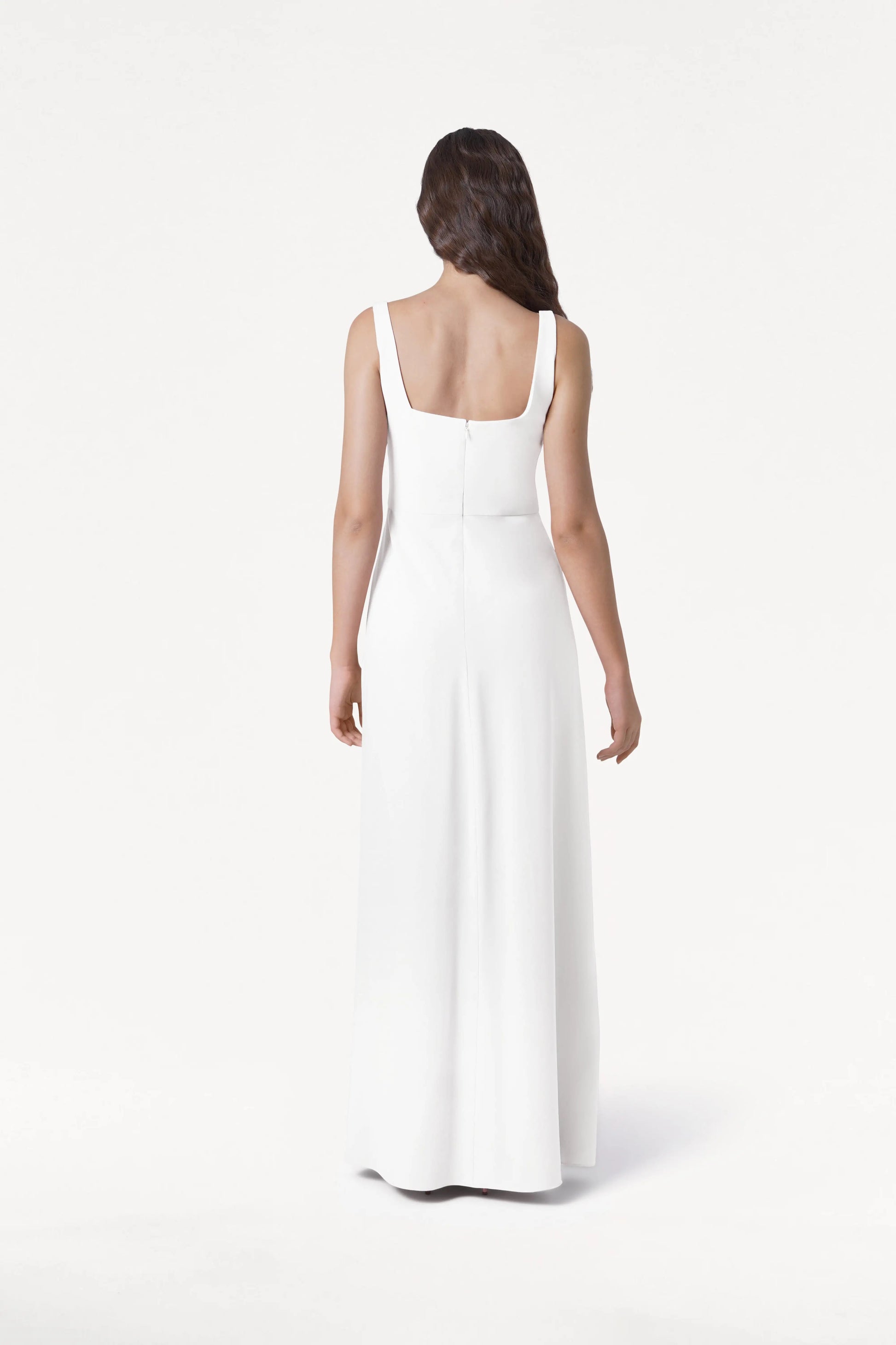 A woman with long, wavy brown hair faces away, wearing the TH&TH Helena Dress in Ivory—a modern, size-inclusive, sleeveless, floor-length bridesmaid dress with a square back and fitted waist—set against a plain white background.
