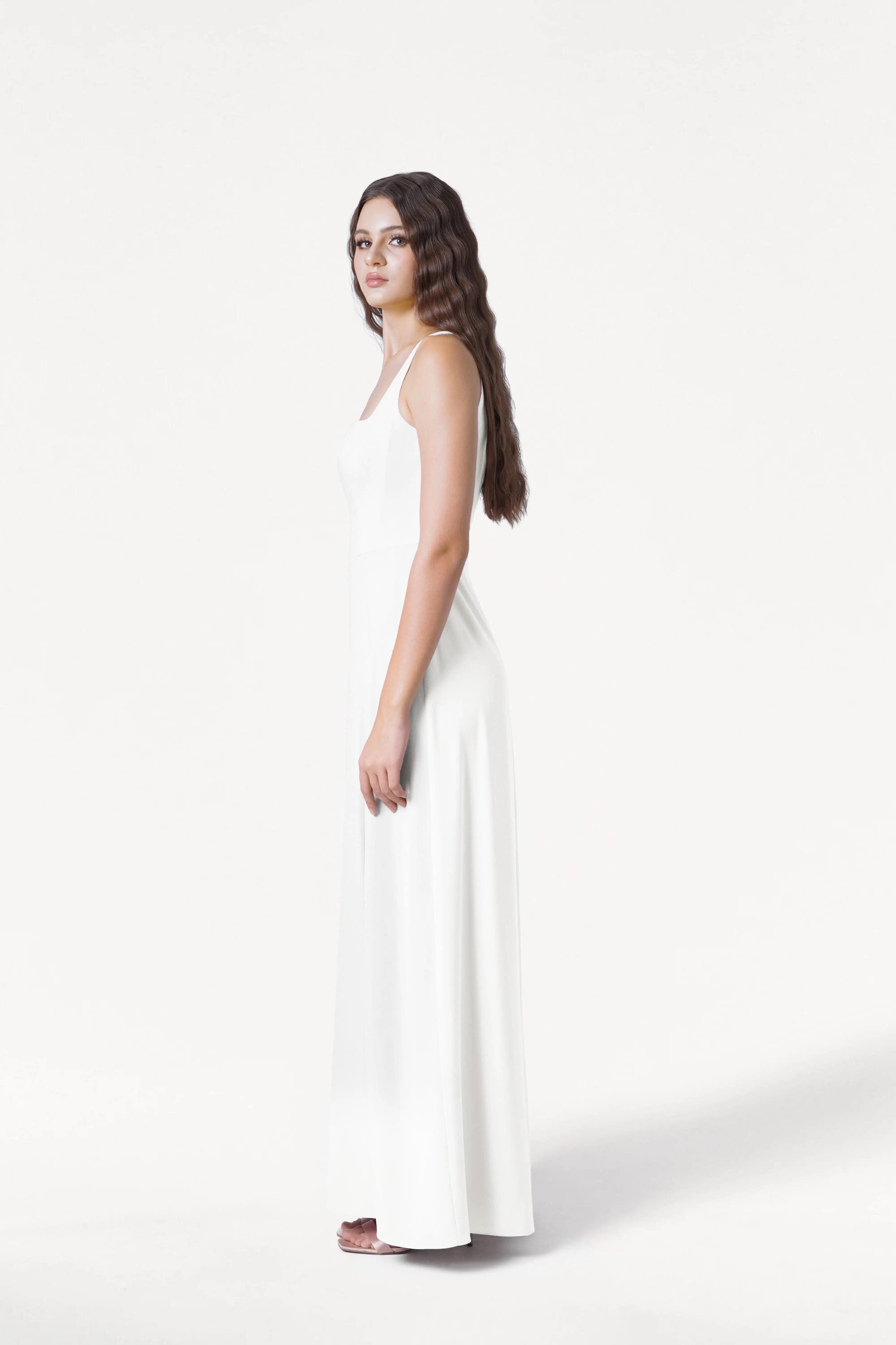 A woman with long wavy hair stands sideways in the TH&TH Helena Dress in Ivory, a sleeveless floor-length modern bridesmaid dress, against a plain white background.
