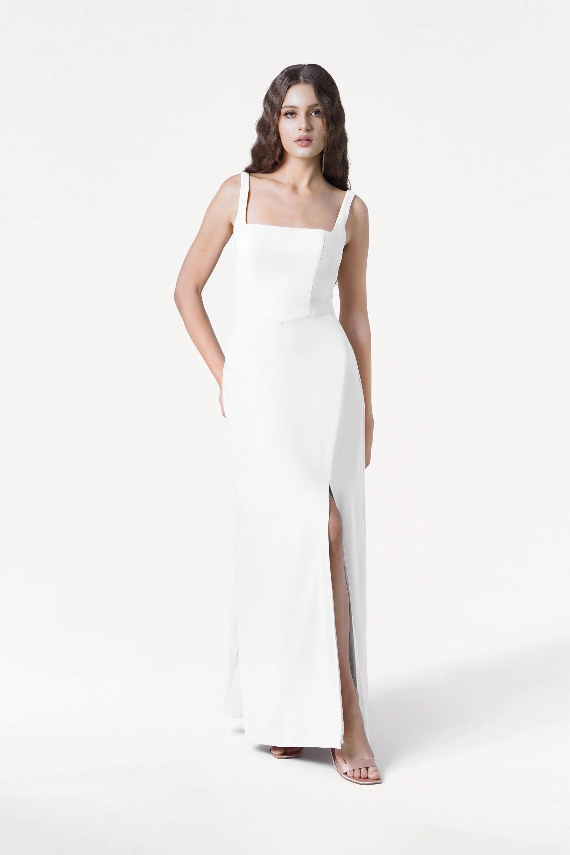 A woman models the TH&TH Helena Dress in Ivory, a modern moss crepe bridesmaid gown with a square neckline and thigh-high slit. She poses against a white background, her wavy brown hair loose, finishing the look with open-toe heels.
