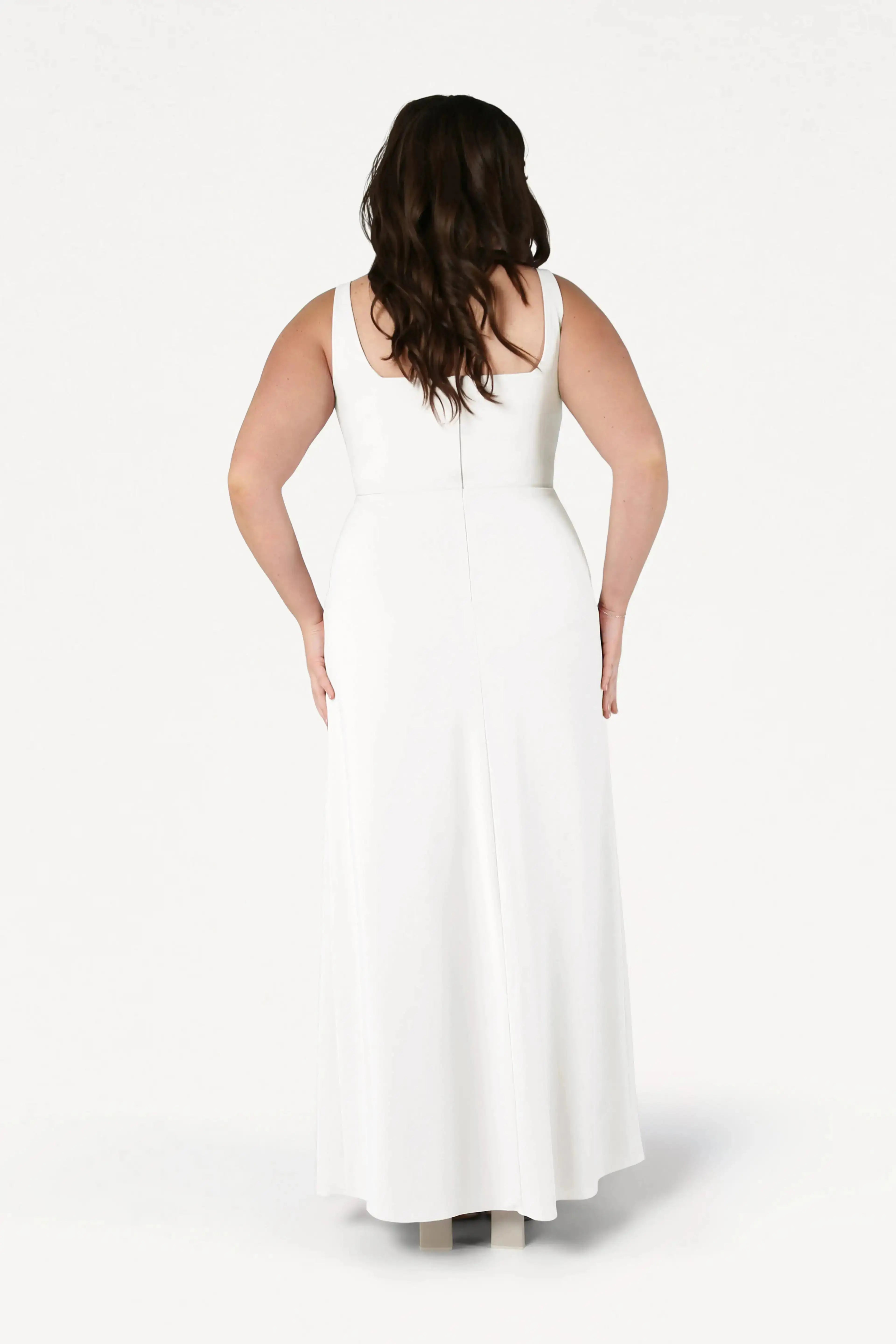 A woman with long brown hair stands facing away, wearing the TH&TH Helena Dress in Ivory—a sleeveless, floor-length, size-inclusive white bridesmaid gown with a fitted waist—set against a plain white background.