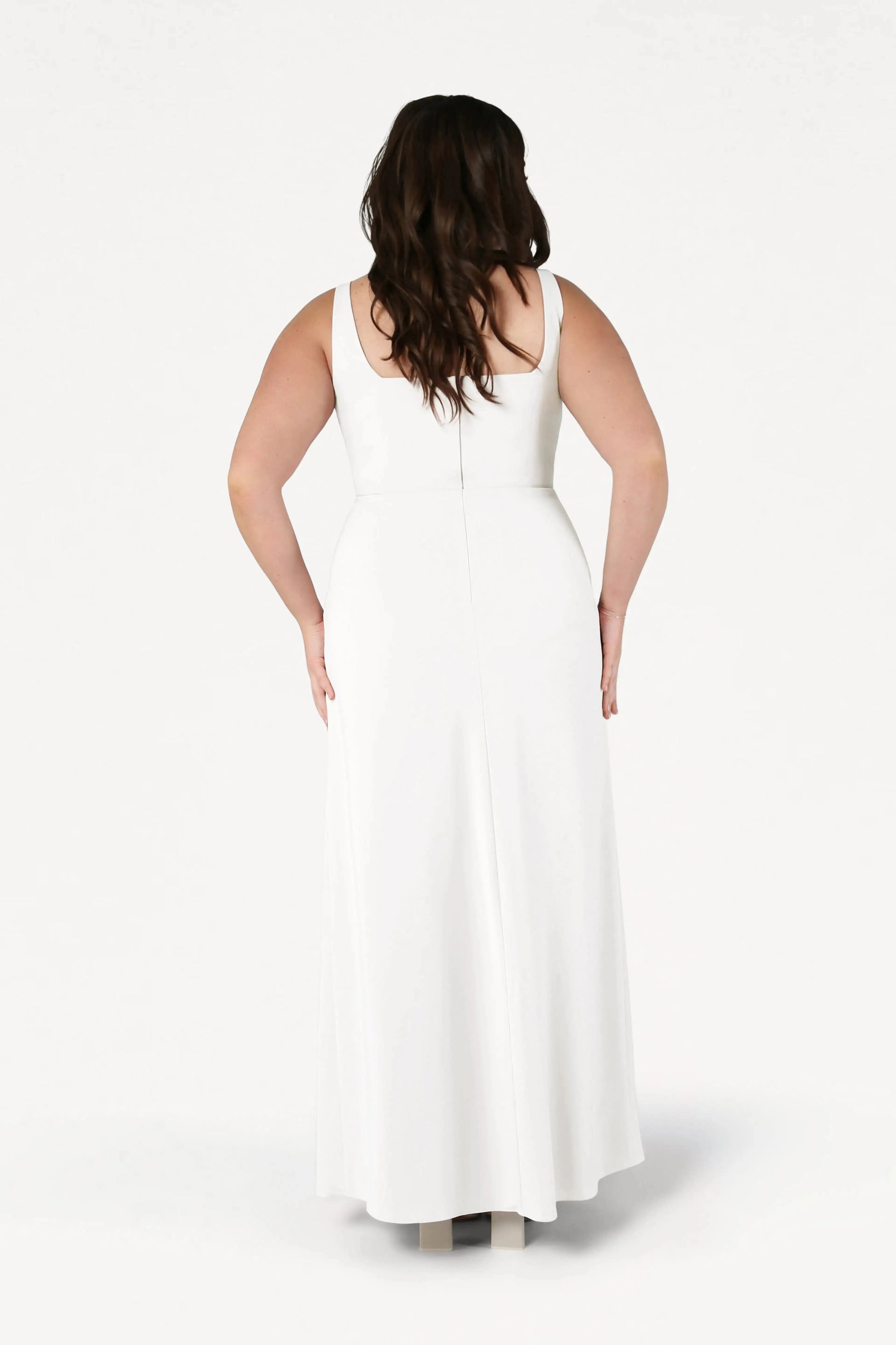 A woman with long brown hair stands facing away, wearing the TH&TH Helena Dress in Ivory—a sleeveless, floor-length, size-inclusive white bridesmaid gown with a fitted waist—set against a plain white background.