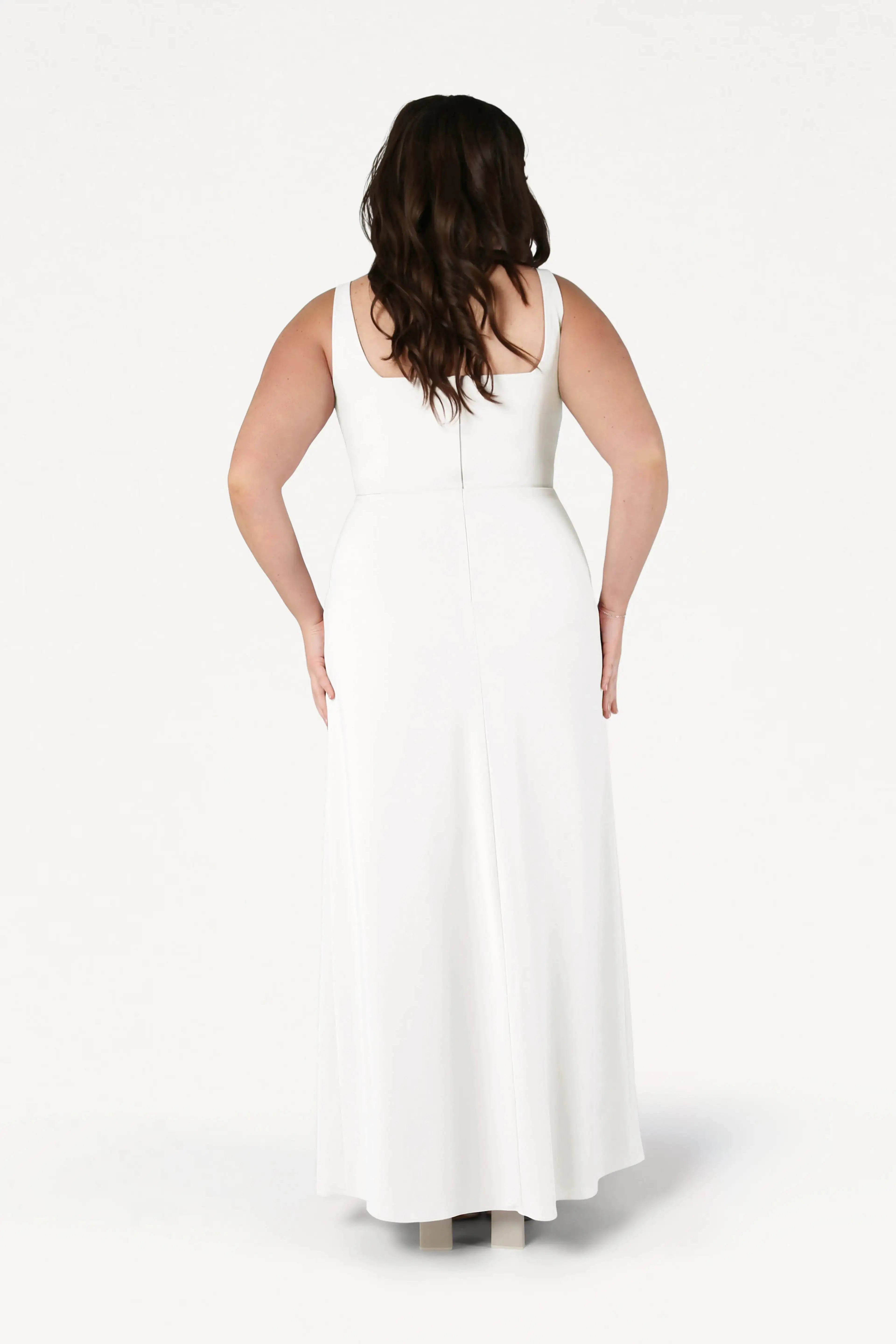 A woman with long brown hair stands facing away, wearing the TH&TH Helena Dress in Ivory—a sleeveless, floor-length, size-inclusive white bridesmaid gown with a fitted waist—set against a plain white background.