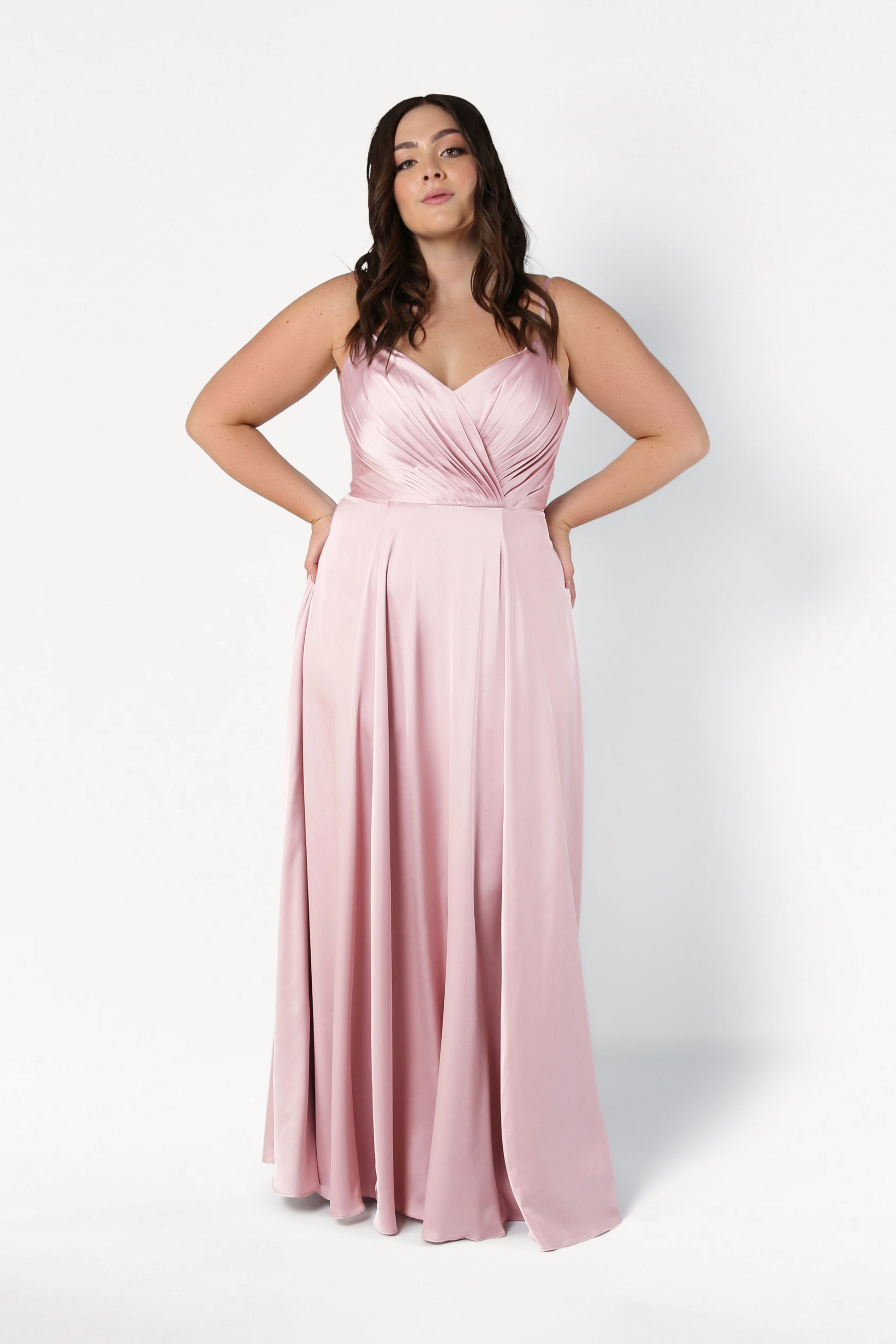 A woman wears the TH&TH Isla Bridesmaid Dress in blush pink satin, featuring a pleated bodice and spaghetti straps, as she stands confidently against a white background with her hands at her hips.